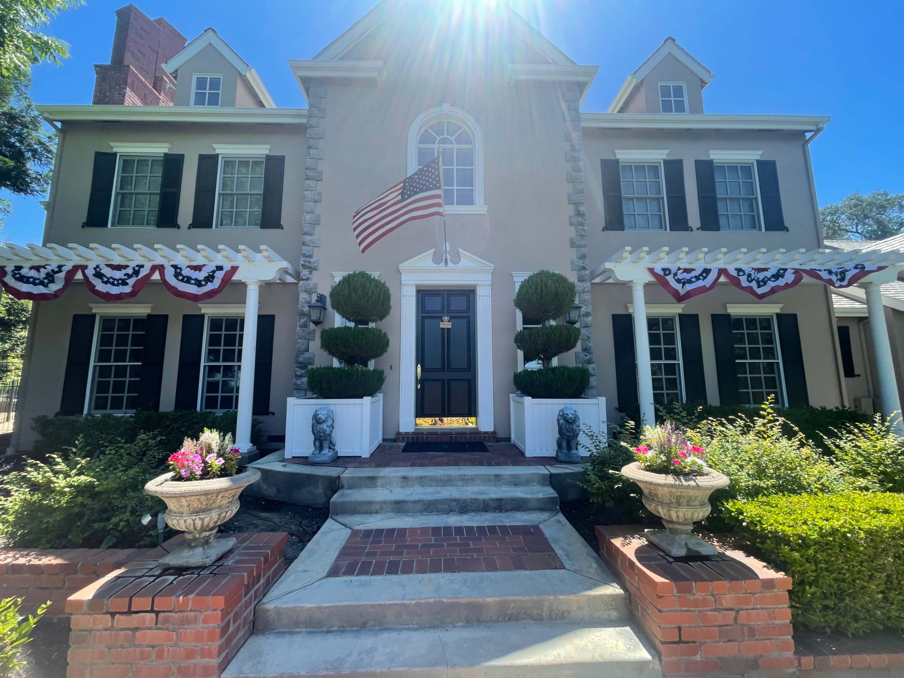 A two-story house with American flags and patriotic bunting displayed on the front porch, surrounded by neatly landscaped gardens and brick walkways.