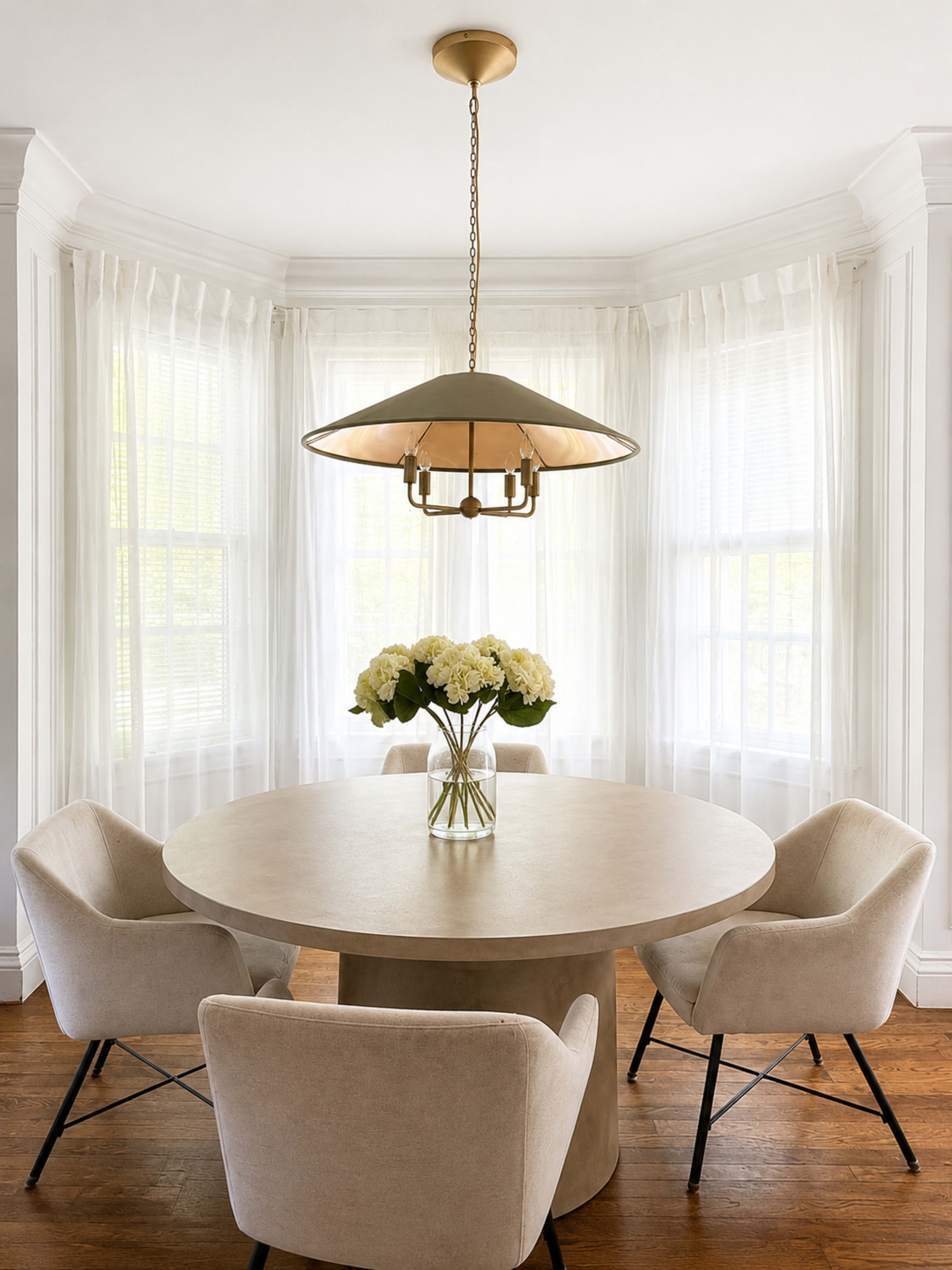 A round table with a vase of white flowers is surrounded by four beige armchairs in a sunlit room with large windows covered by sheer curtains.