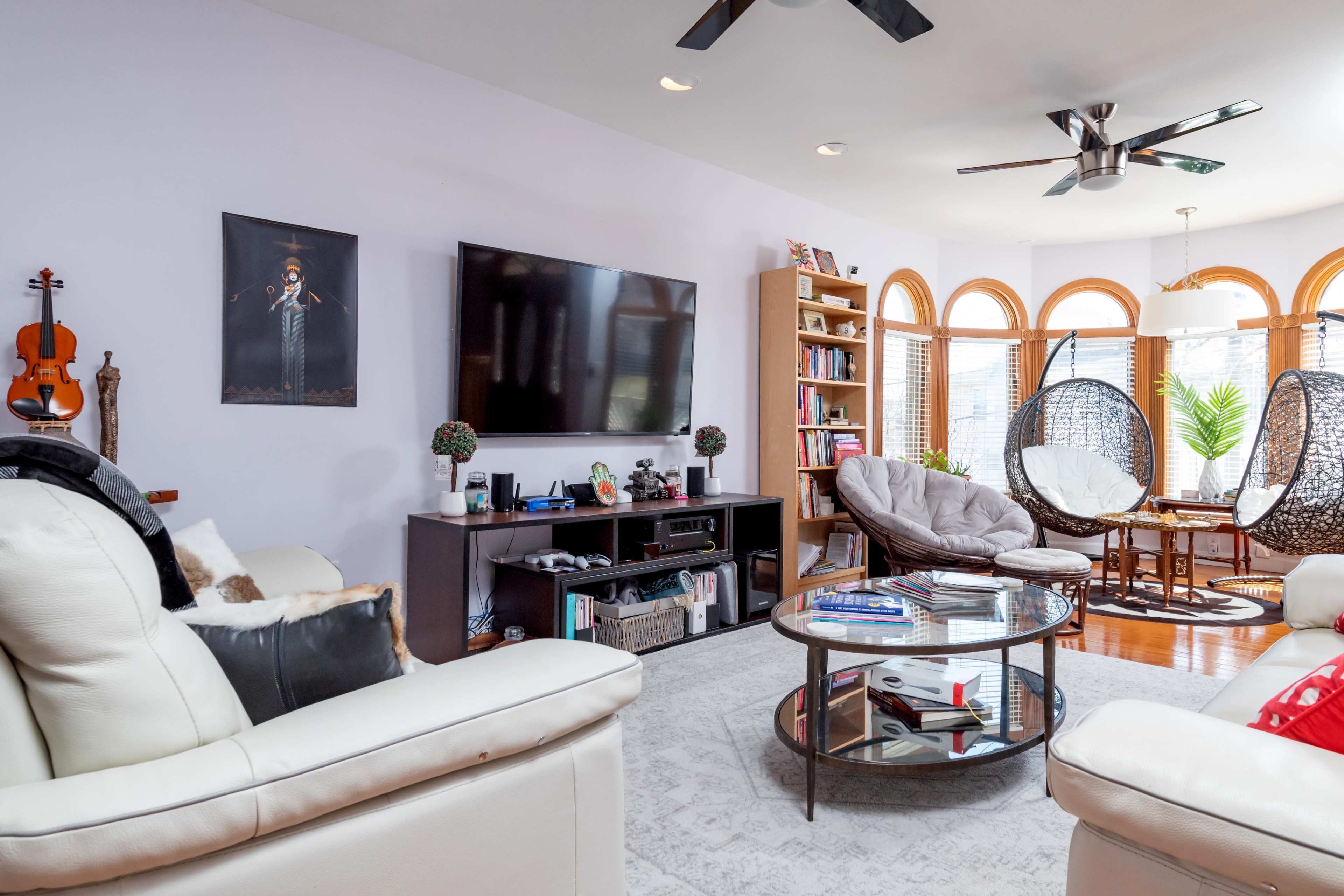 A modern living room featuring two hanging chairs, a large television, a bookshelf, and a coffee table, with large windows allowing natural light to fill the space.