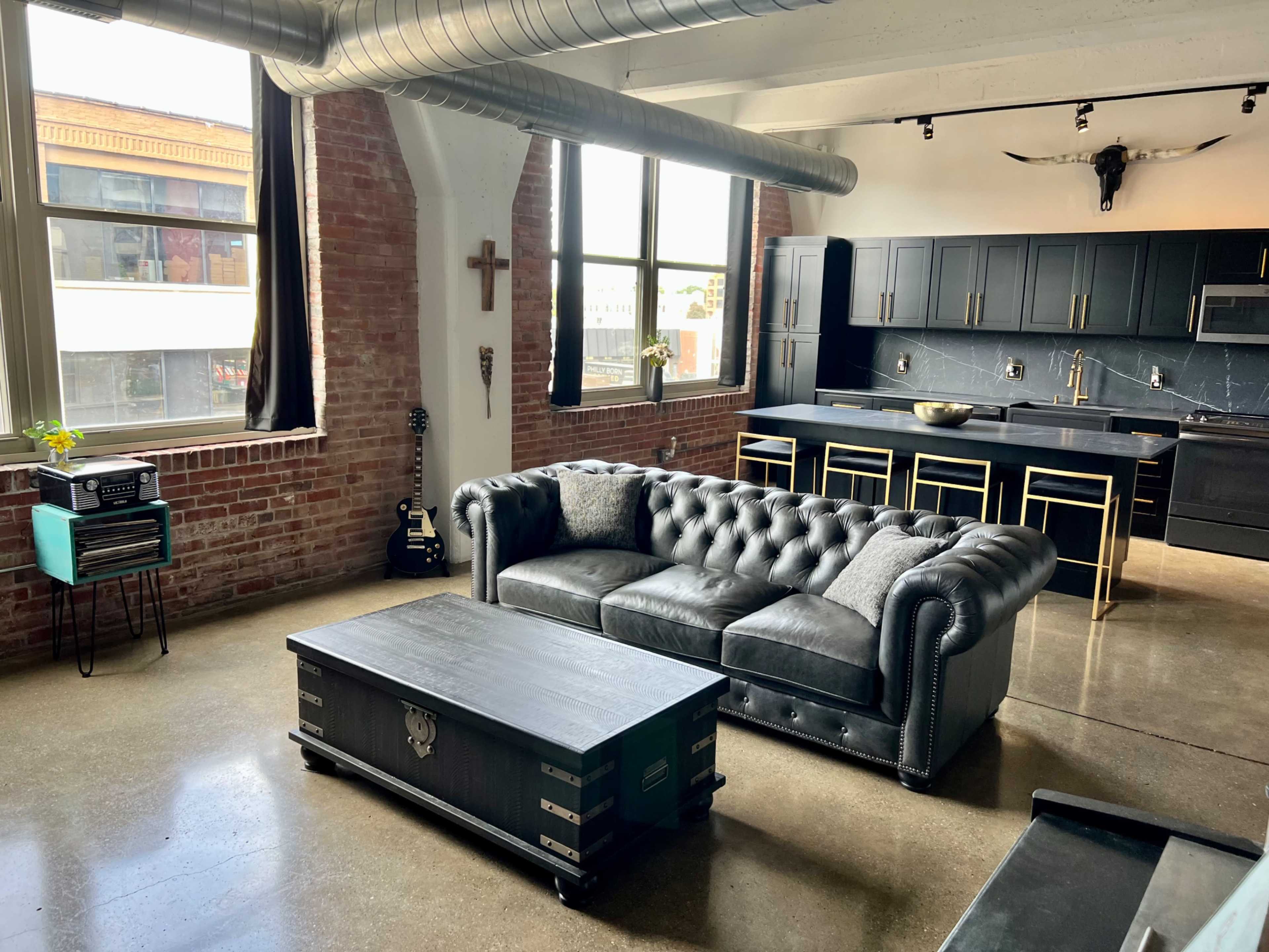 A modern industrial-style living space with a black leather chesterfield sofa, a coffee table, and a kitchen area featuring dark cabinetry and metallic accents.