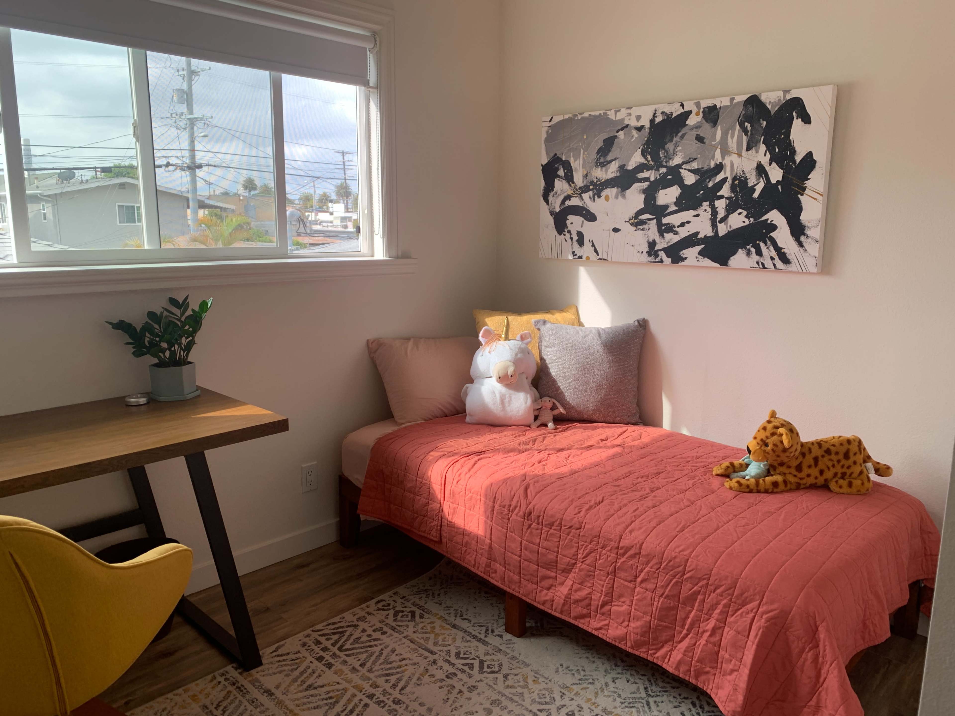 A small room with a single bed covered in a pink quilt, a wooden desk with a green plant, and a large piece of abstract artwork on the wall.