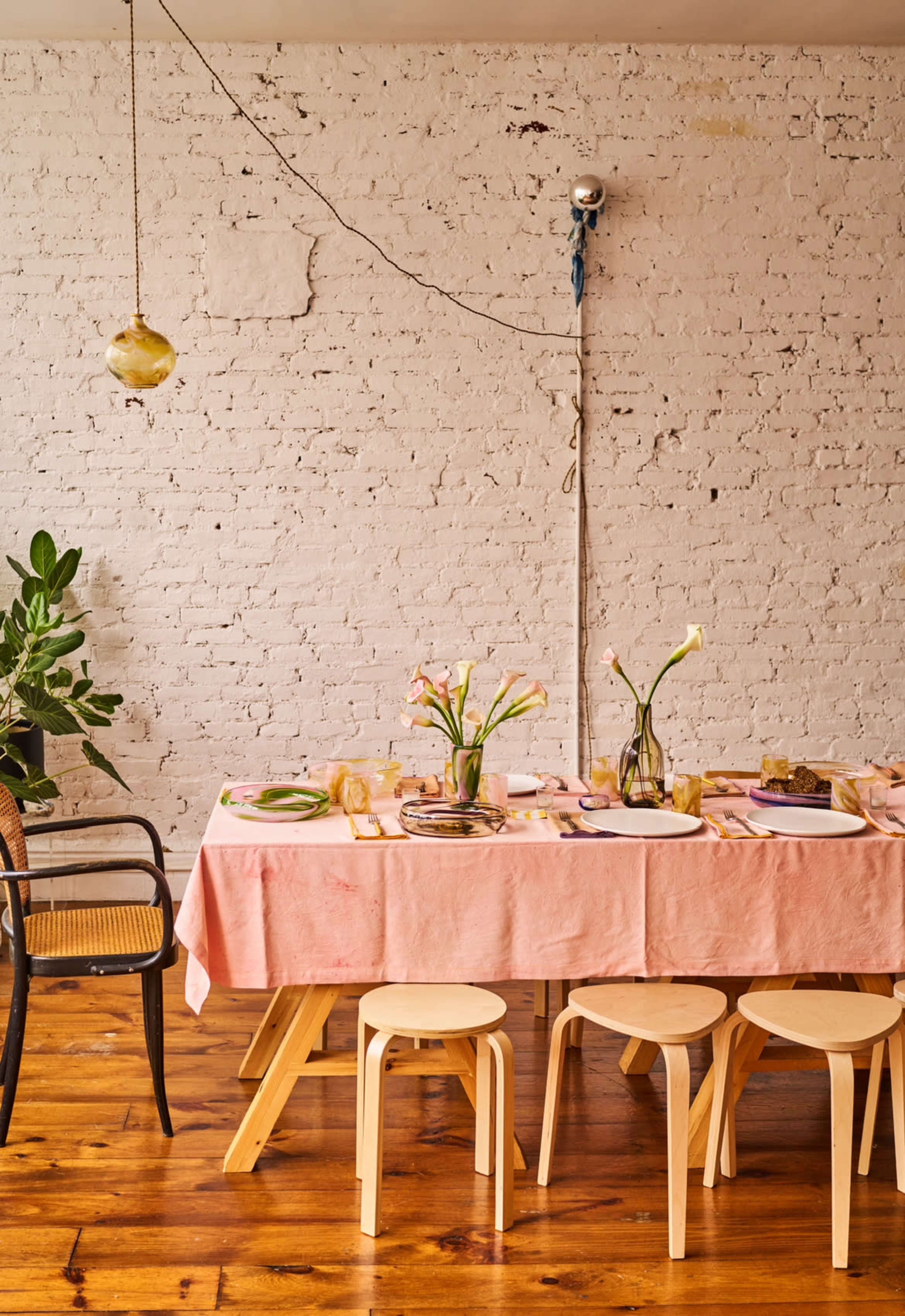 A dining table with a pink tablecloth is set with plates and cutlery, surrounded by wooden stools, against a white brick wall.