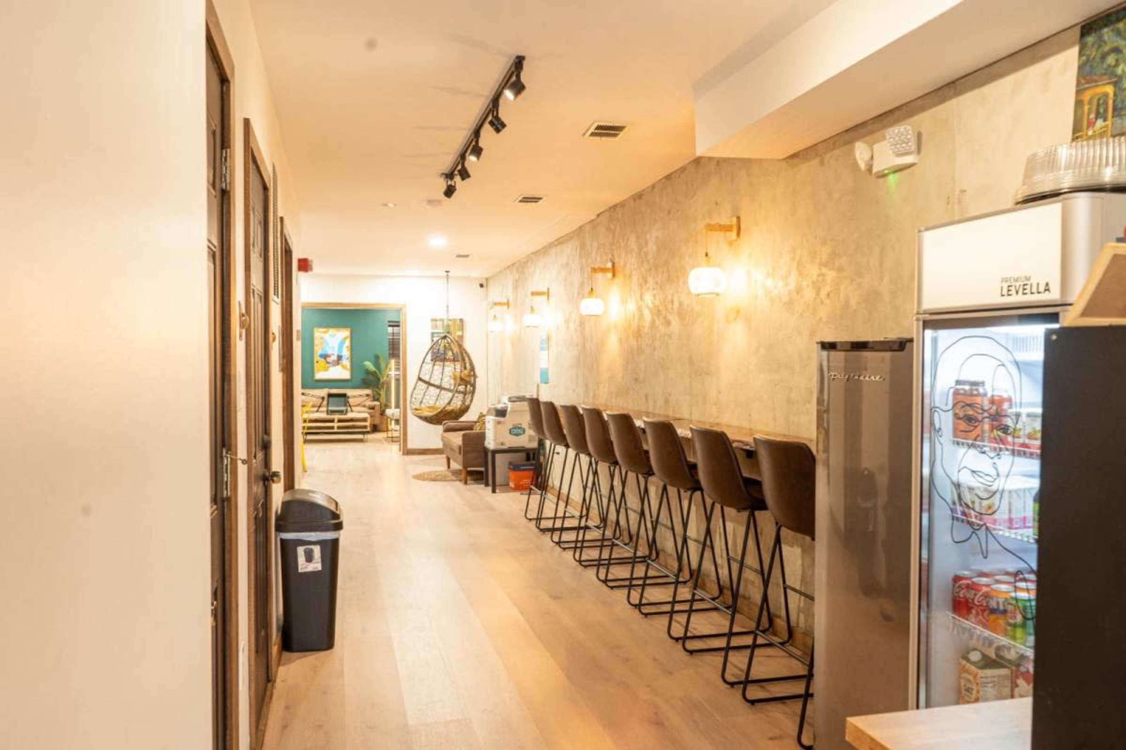 The image shows a modern hallway featuring a row of barstools, a hanging chair, and a refrigerator, with warm lighting illuminating the walls.
