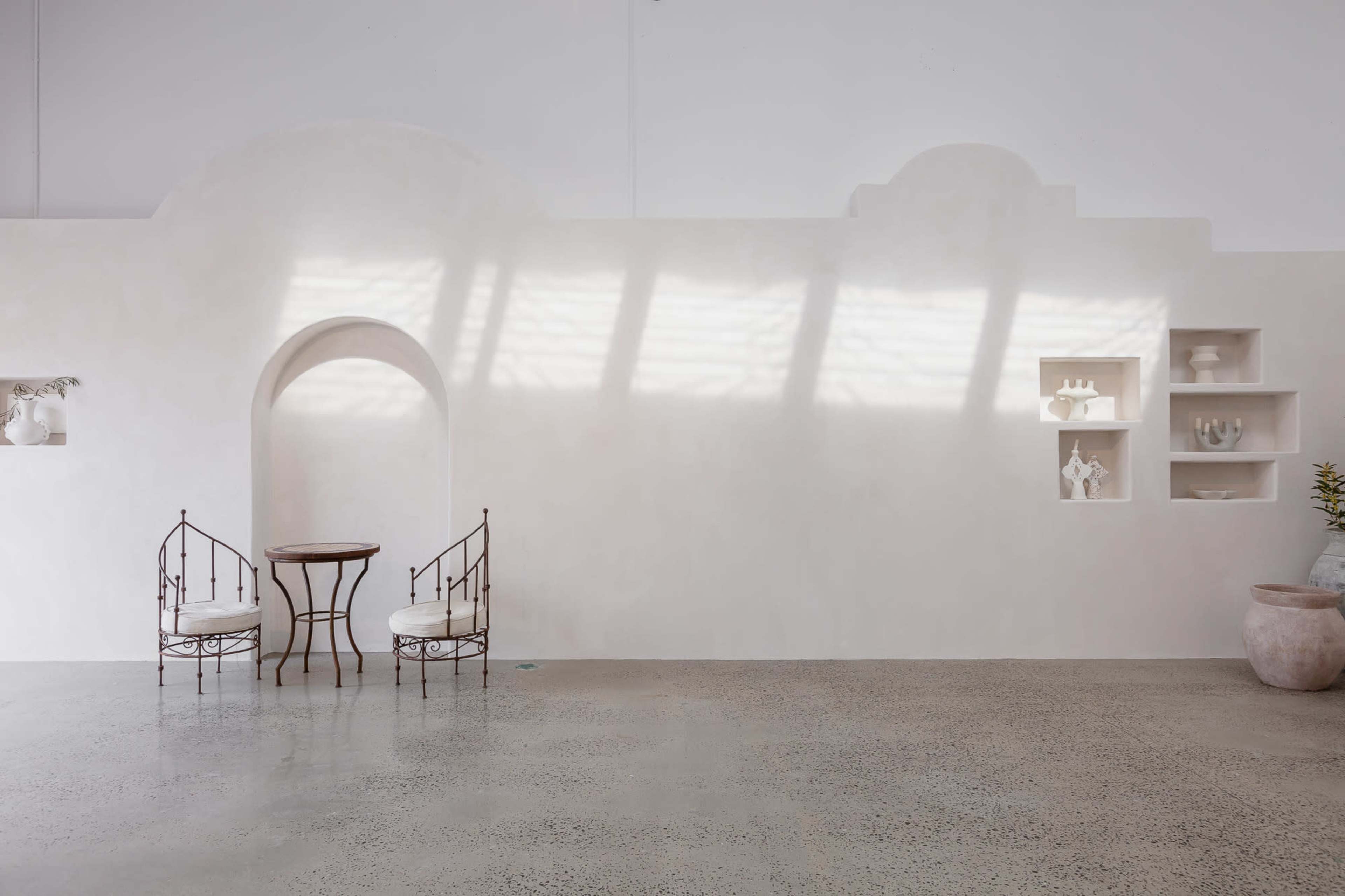 A minimalistic interior space with a light-colored wall, a small round table, two chairs, and built-in shelves displaying decorative objects.