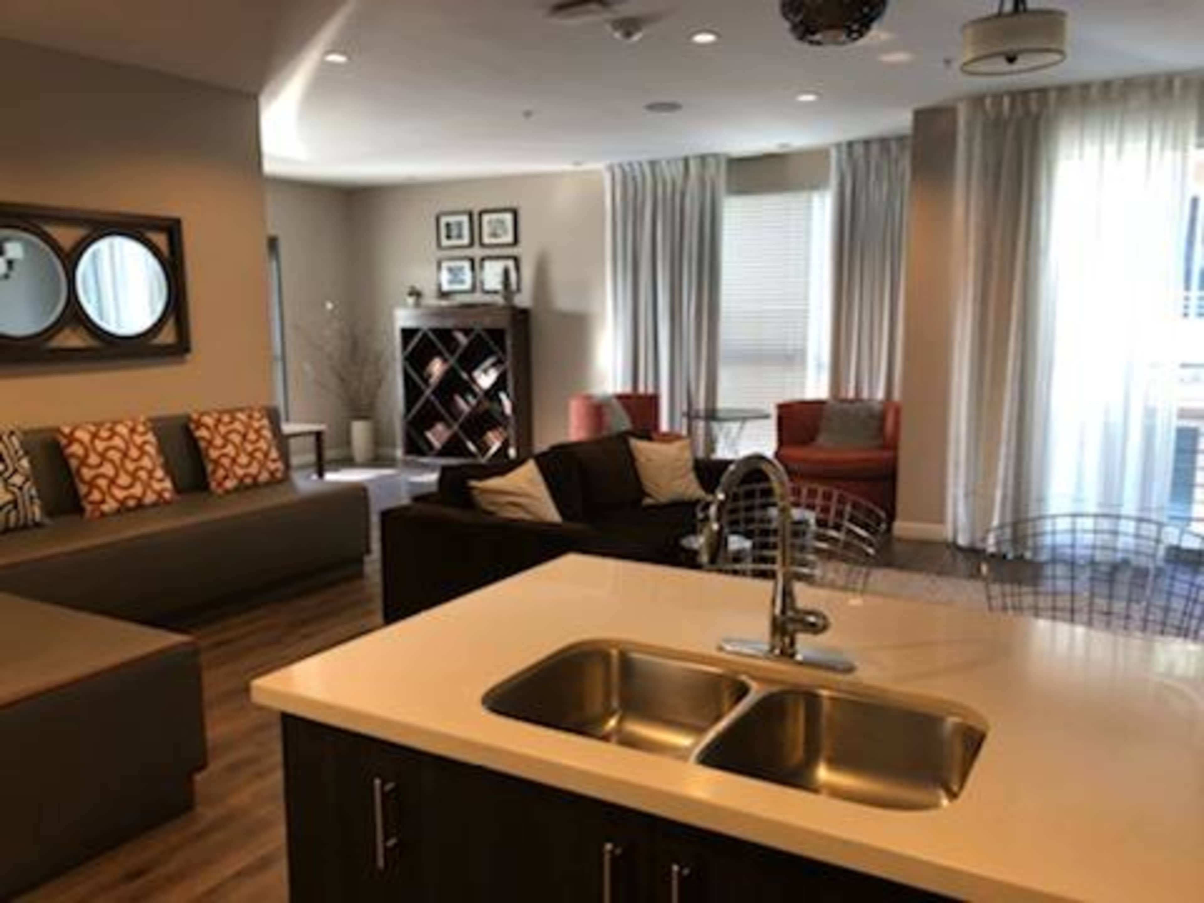 A modern living area with a kitchen island featuring a double sink, seating areas with couches and chairs, and large windows covered by curtains.