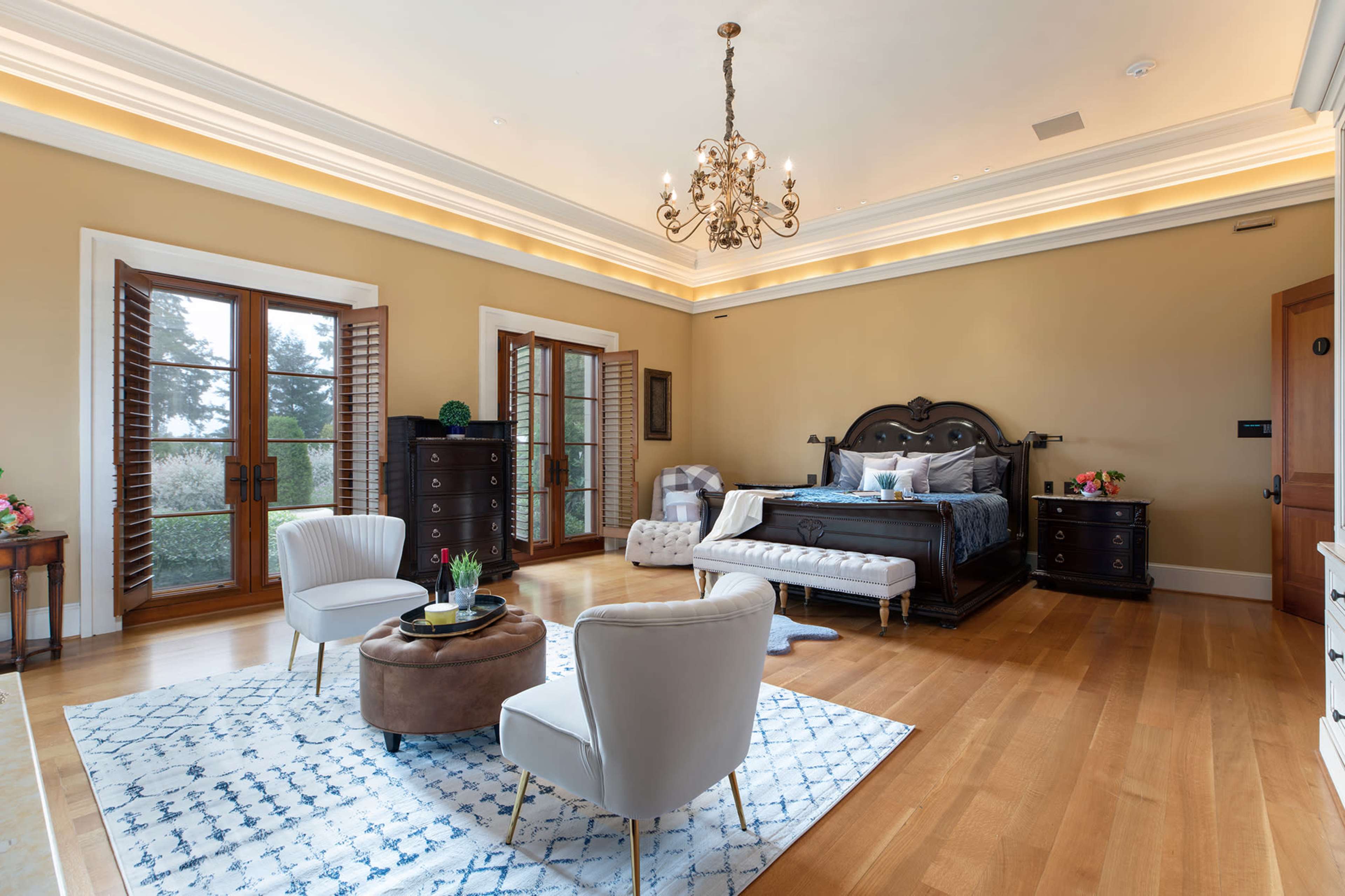 A spacious bedroom featuring a large ornate bed, two upholstered chairs, a small round coffee table, and windows with wooden shutters.