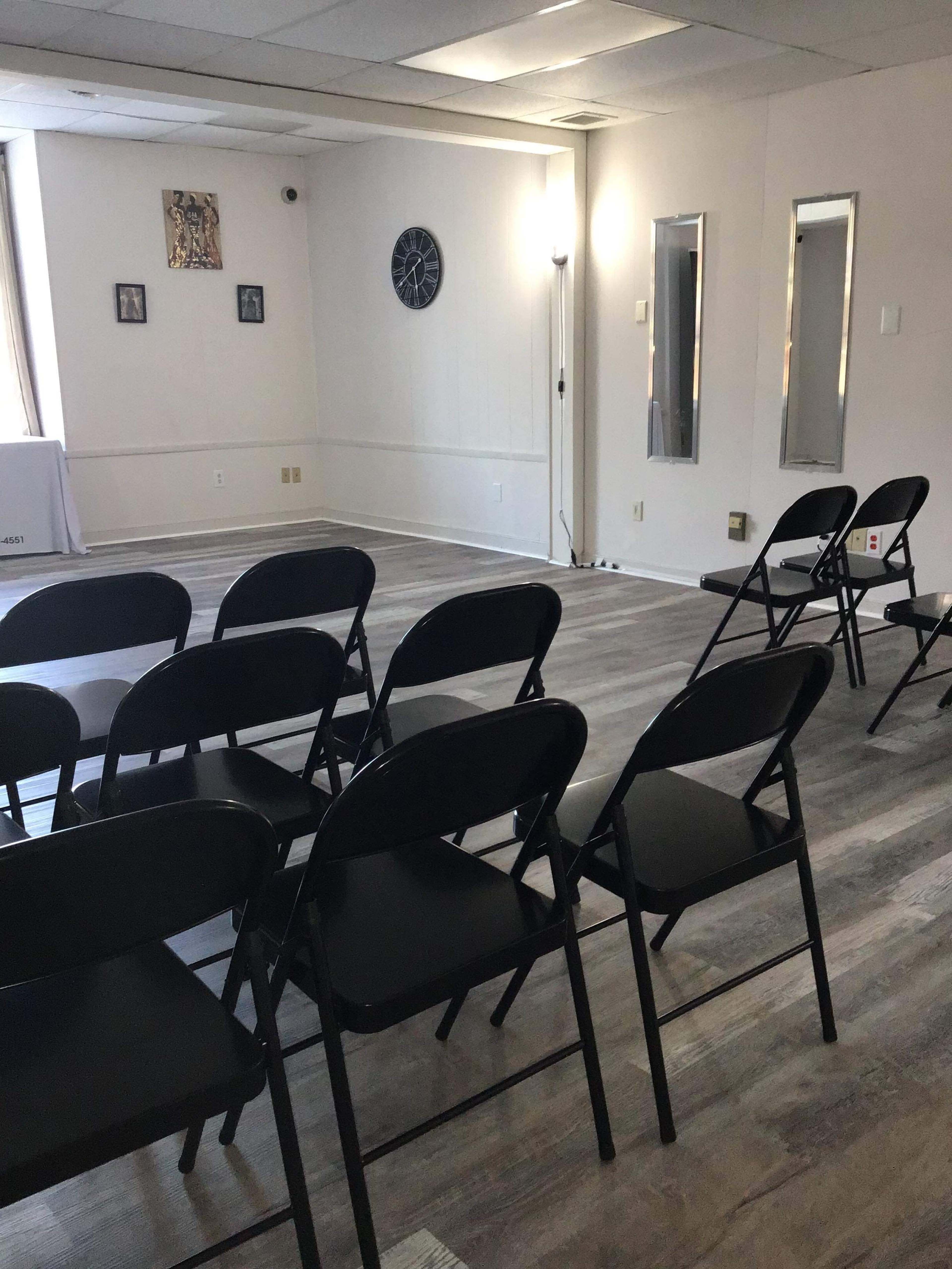 A row of black folding chairs is arranged in a light-filled room featuring mirrors and a wall clock.