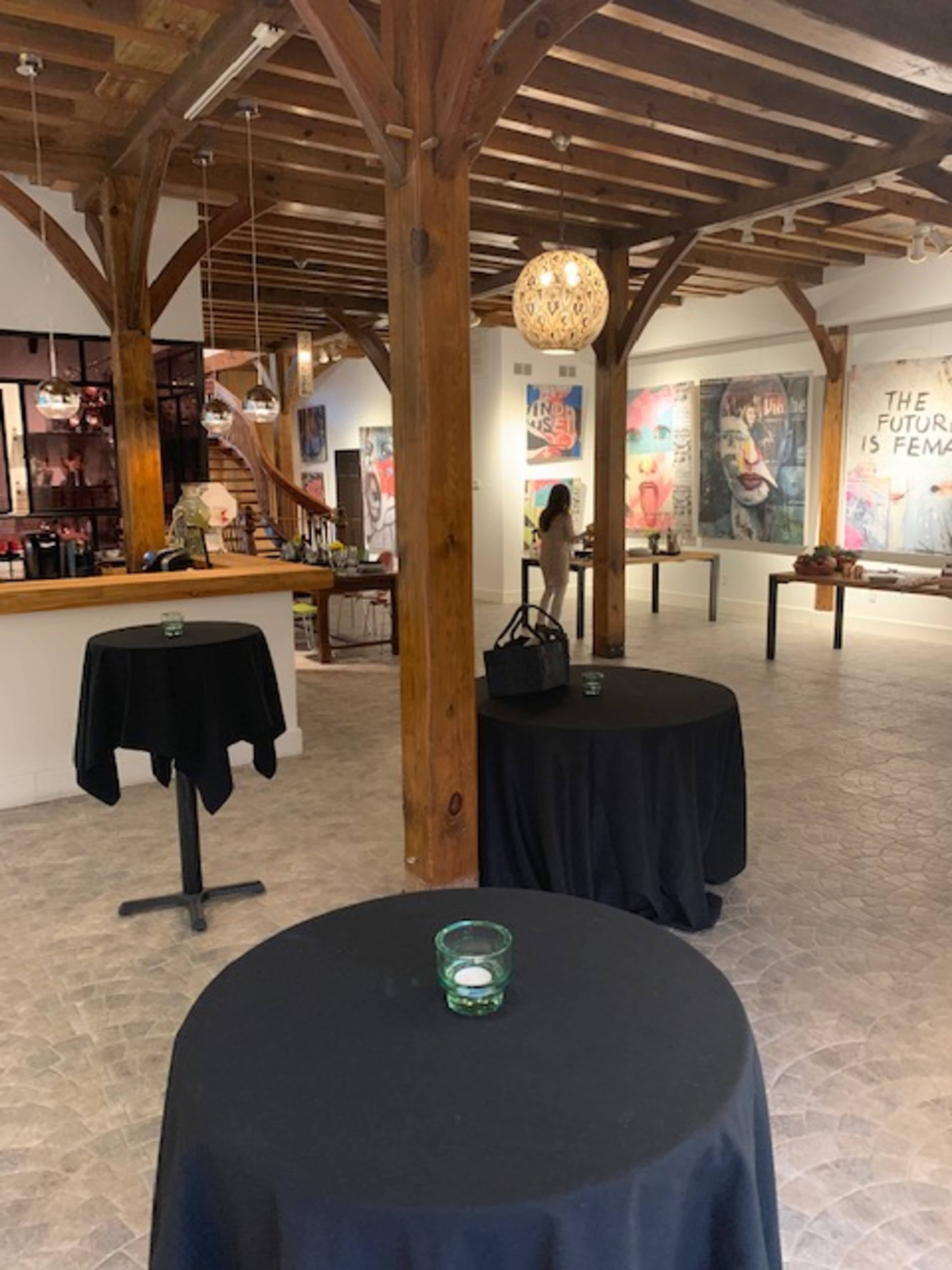 An art gallery with wooden beams, several black tables, and artworks displayed on the walls.