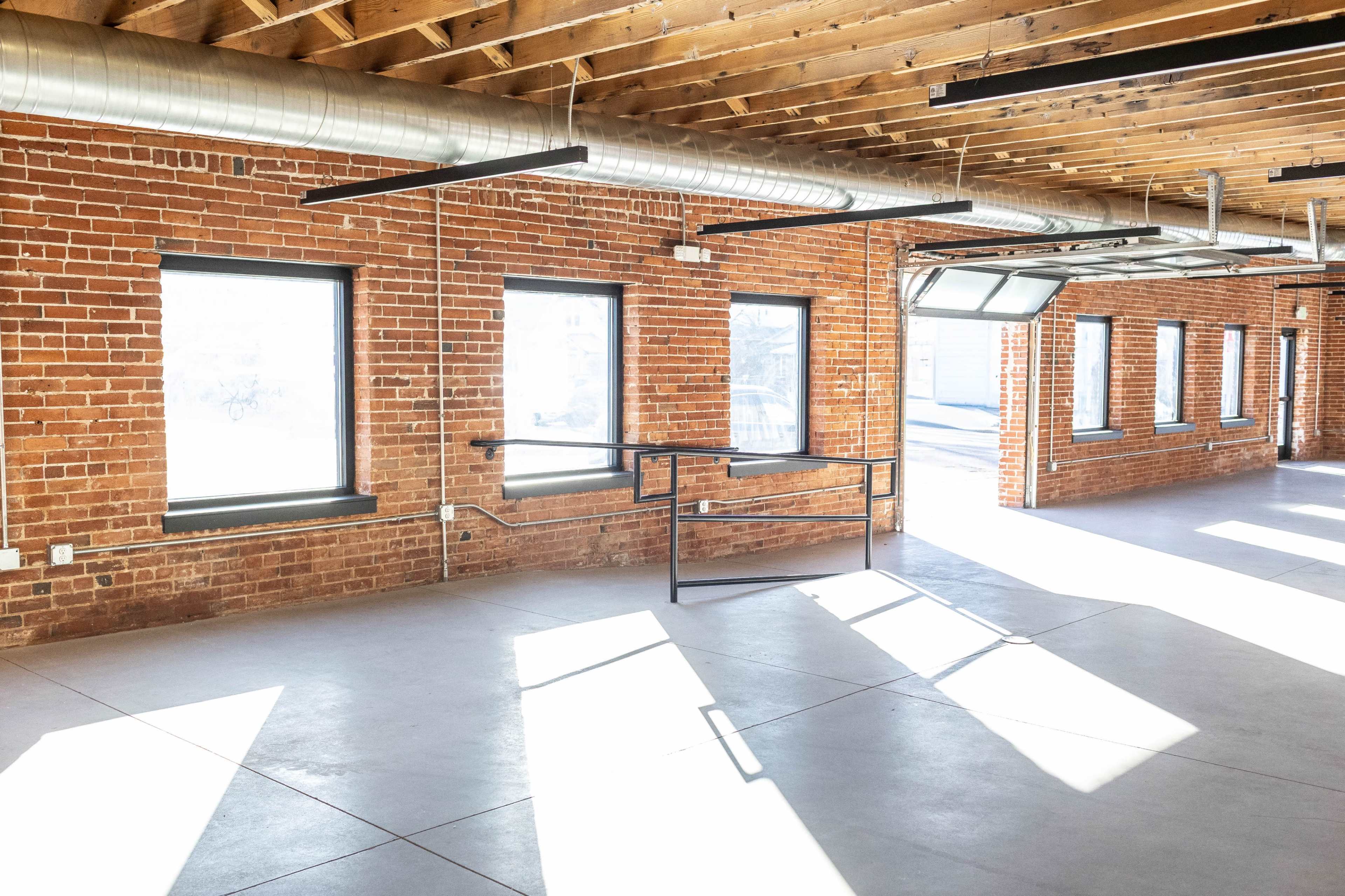 The image shows a spacious room with exposed brick walls, large windows, a metal railing, and sunlight casting shadows on the concrete floor.
