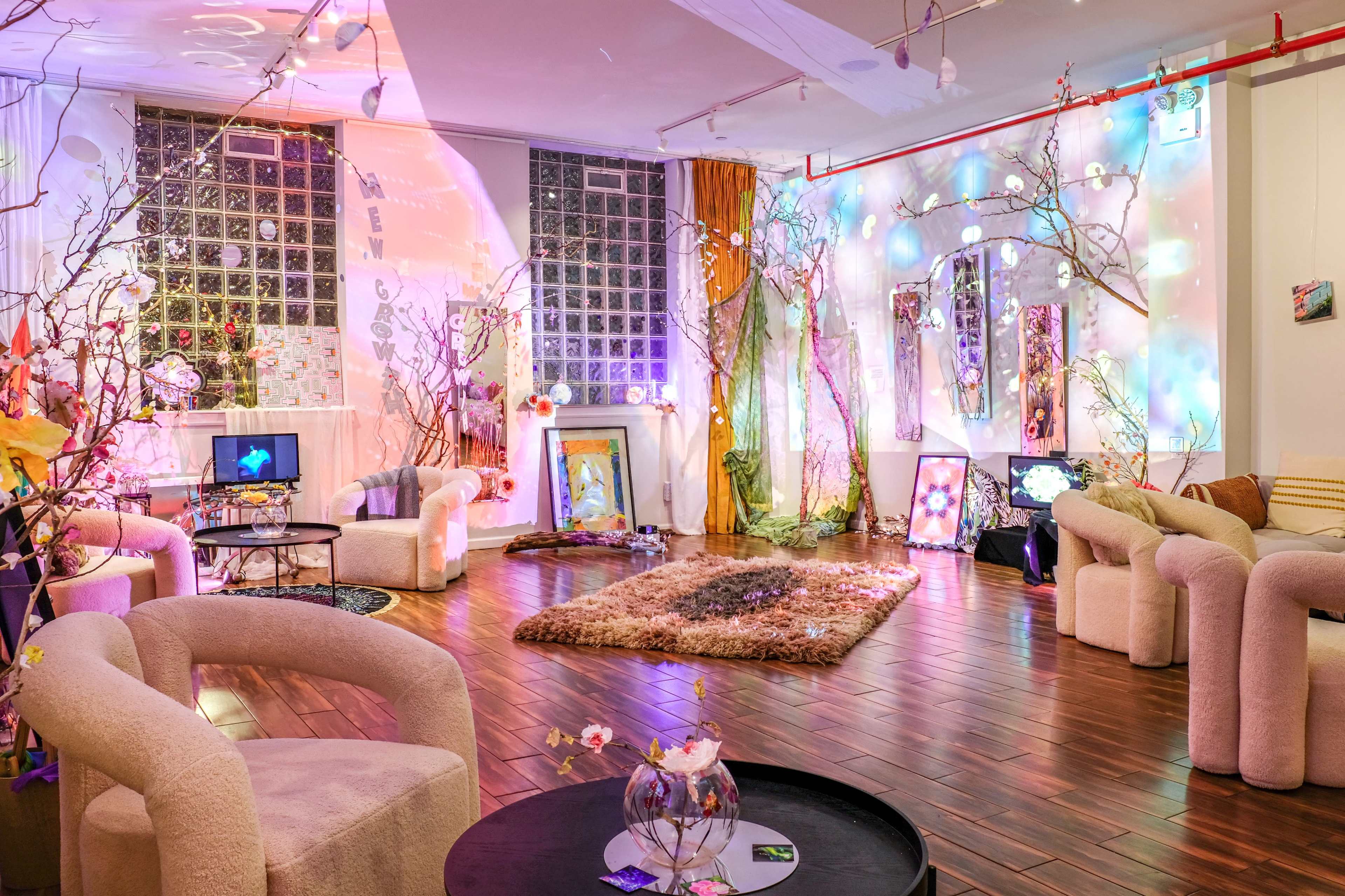 The image shows a brightly lit interior space featuring modern, circular chairs, decorative branches, soft rugs, and colorful lighting effects on the walls.