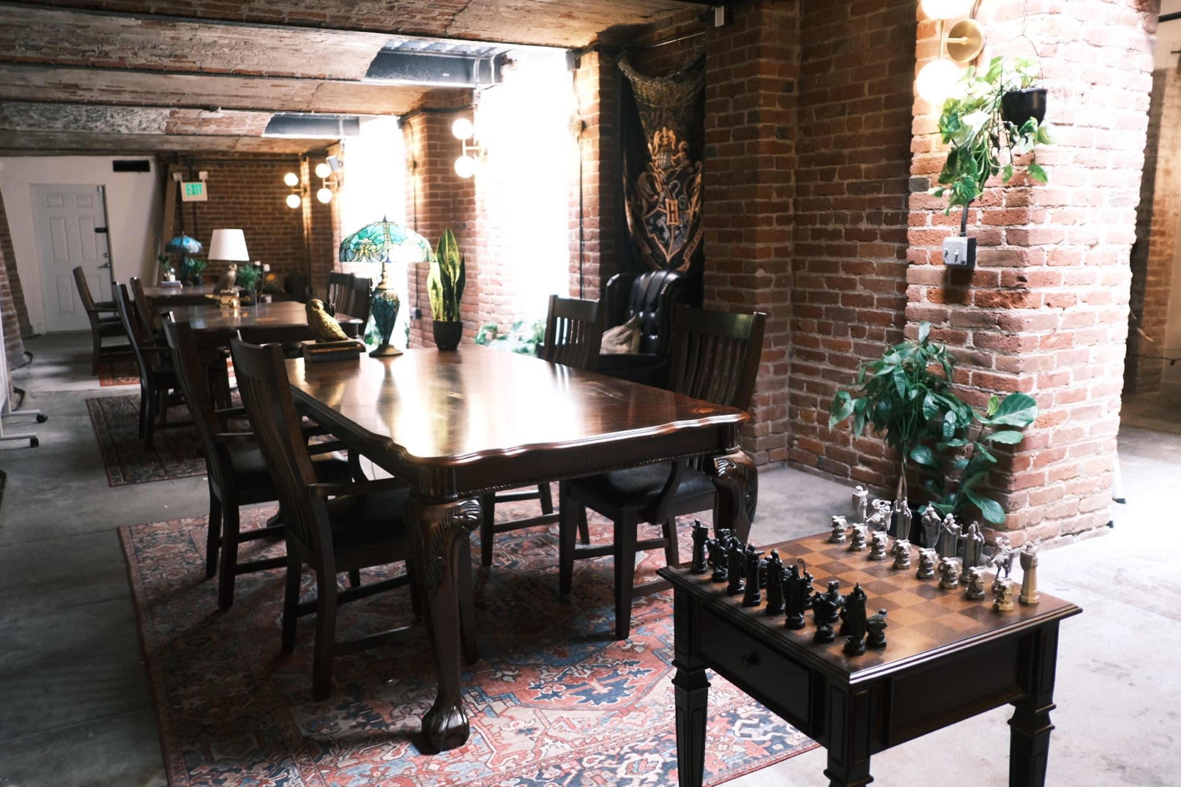 A large dining table is set in a room with exposed brick walls and lighting, accompanied by a chess set on a smaller table.
