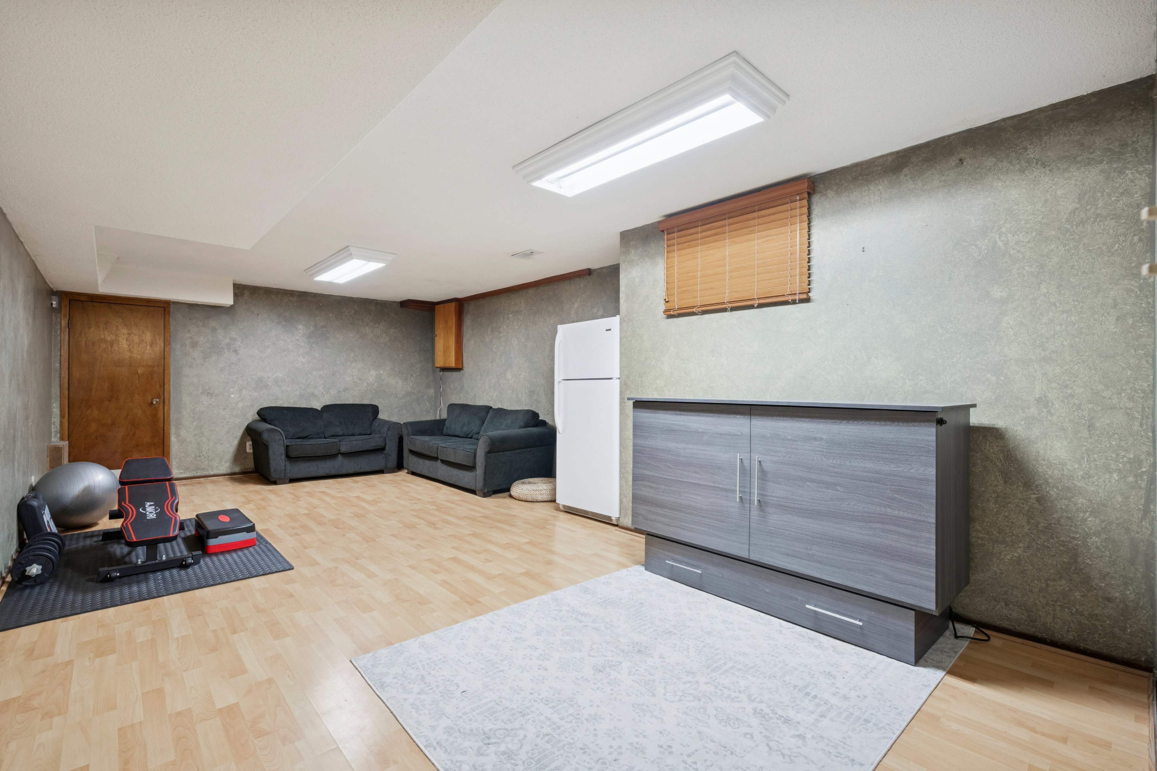 The image shows a sparsely furnished basement with a light-colored wooden floor, a gray couch, a white refrigerator, and a small area rug.