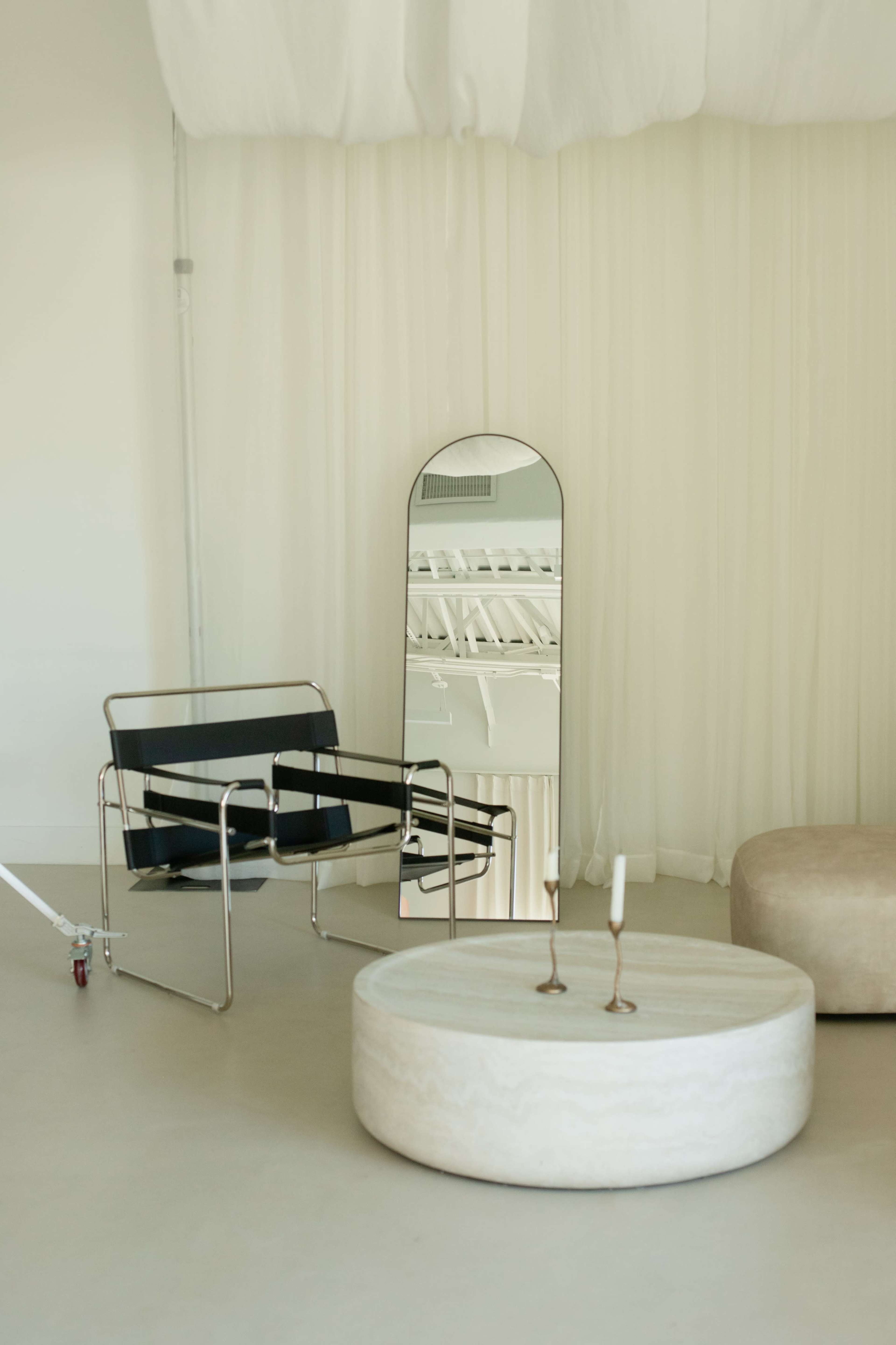 The image features a modern interior with a black chair, a round marble table, and a tall mirror reflecting an airy, minimalist space with soft drapery.