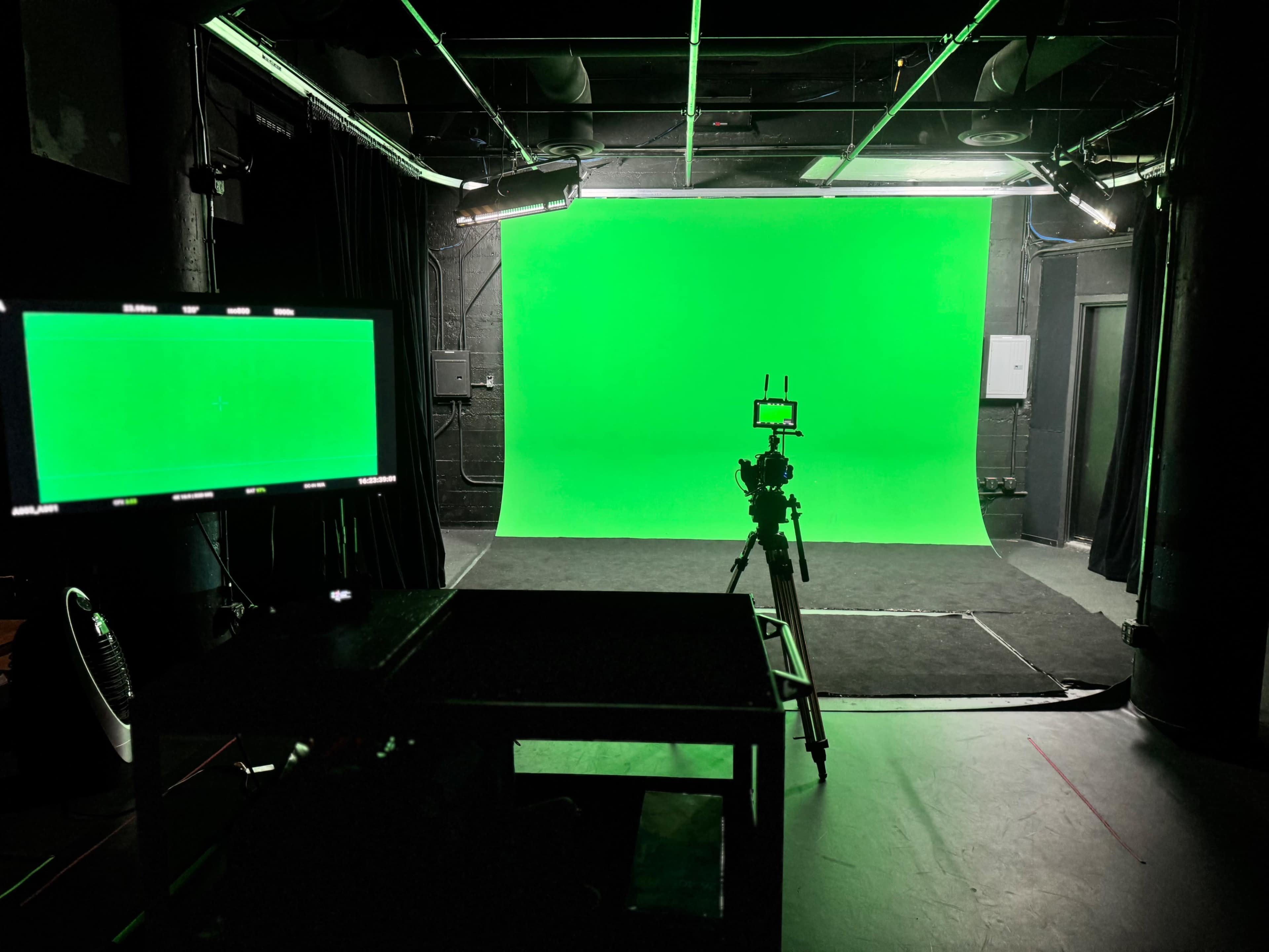 The image shows a film studio with a green screen backdrop, a camera on a tripod, and a monitor on a table.