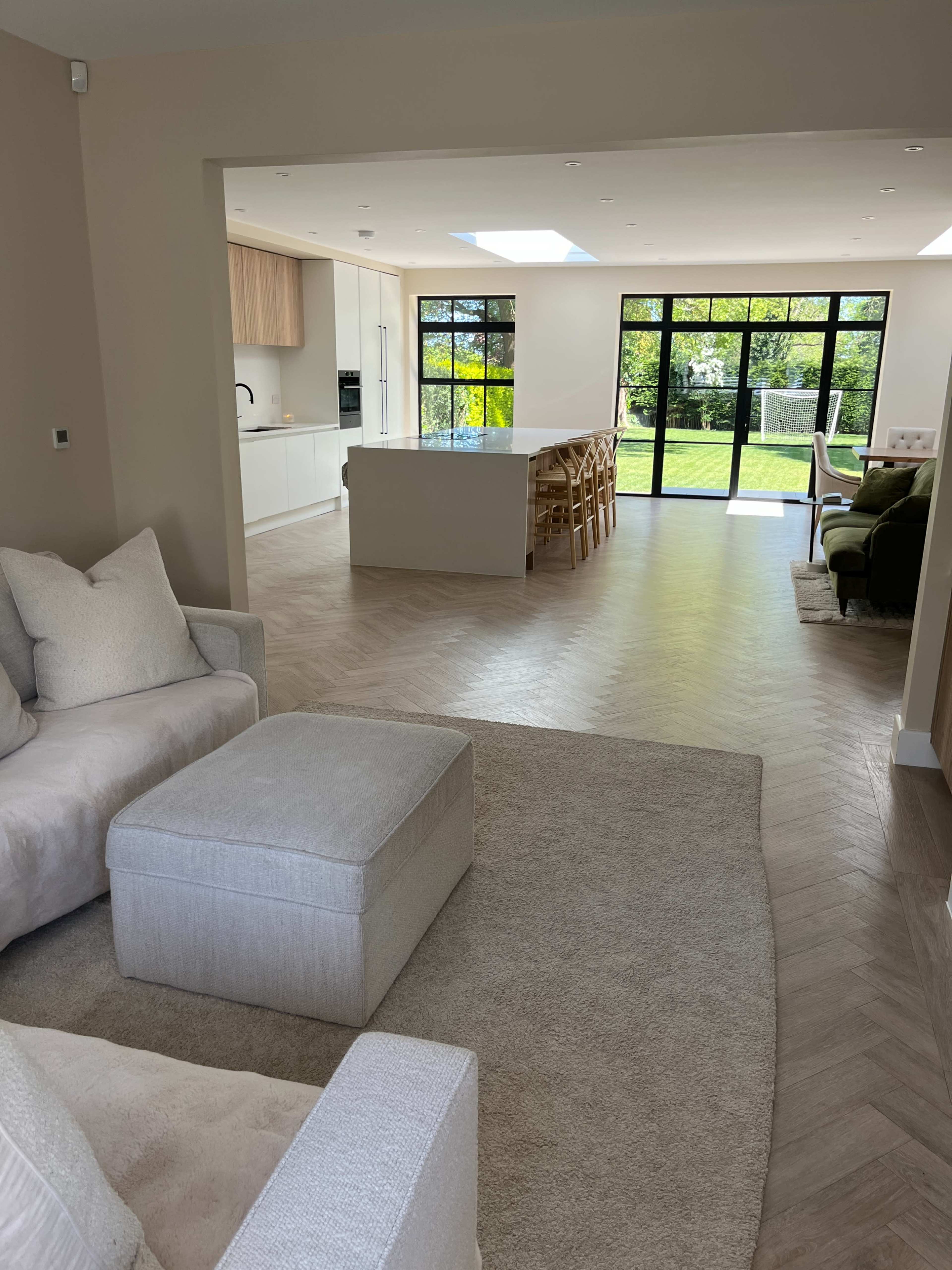 The image shows a modern, open-concept living space with light-colored furniture, a dining area, and large windows overlooking a green outdoor area.