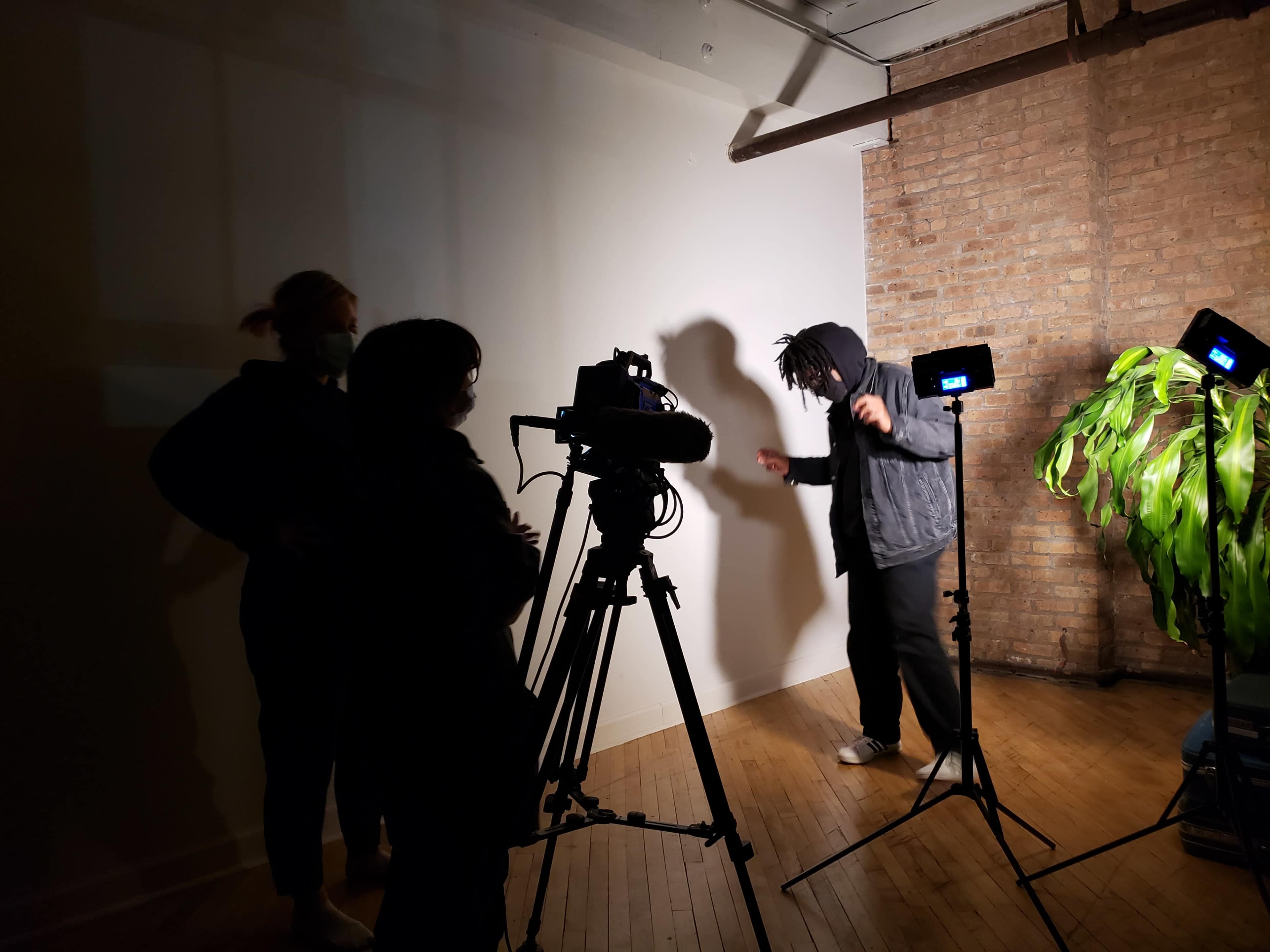 A person in a hoodie performs in front of a camera and two lights, while two others observe in a studio setting.