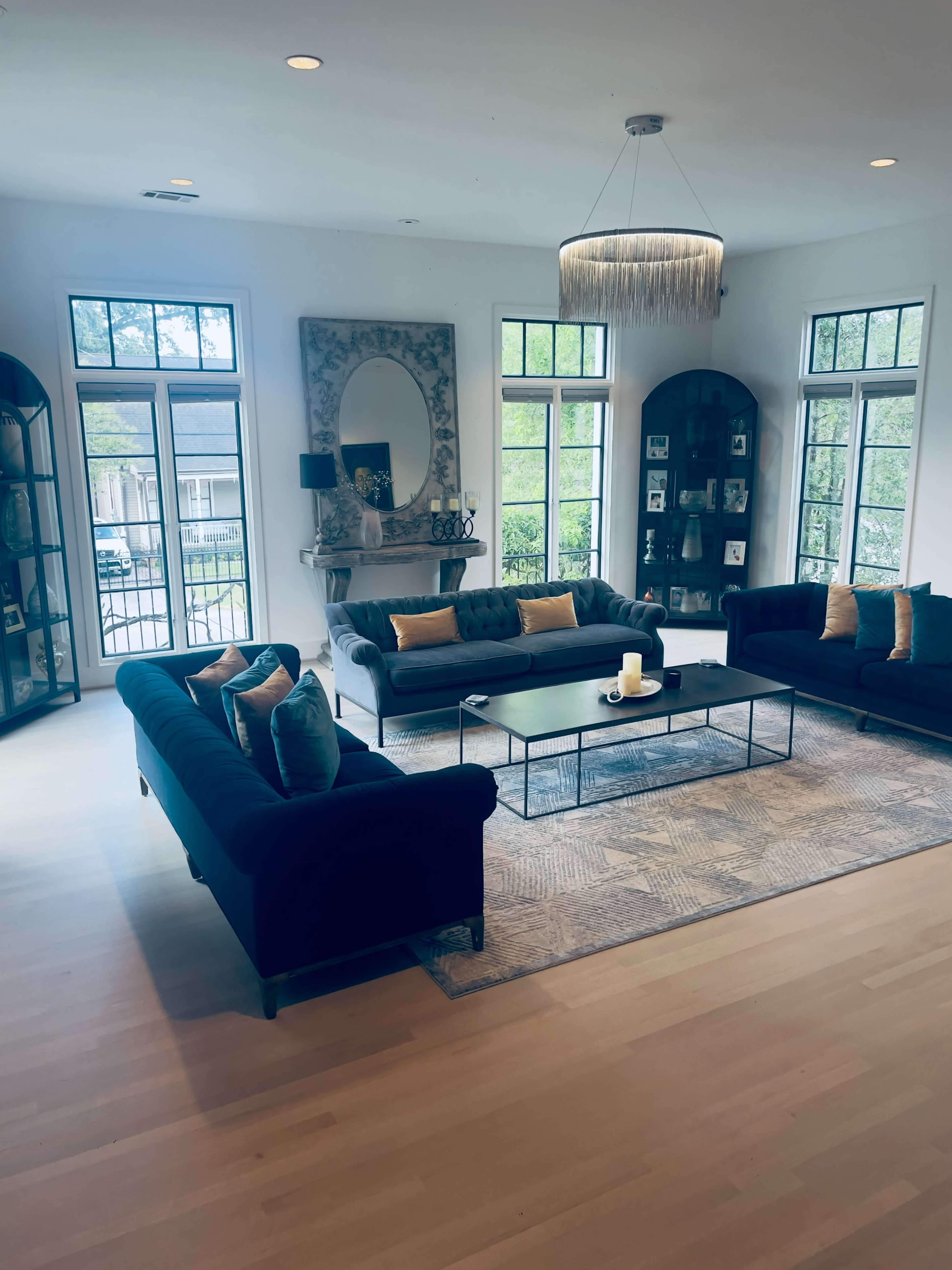 The image shows a spacious living room with large windows, a coffee table in the center, and two black sofas arranged around it.