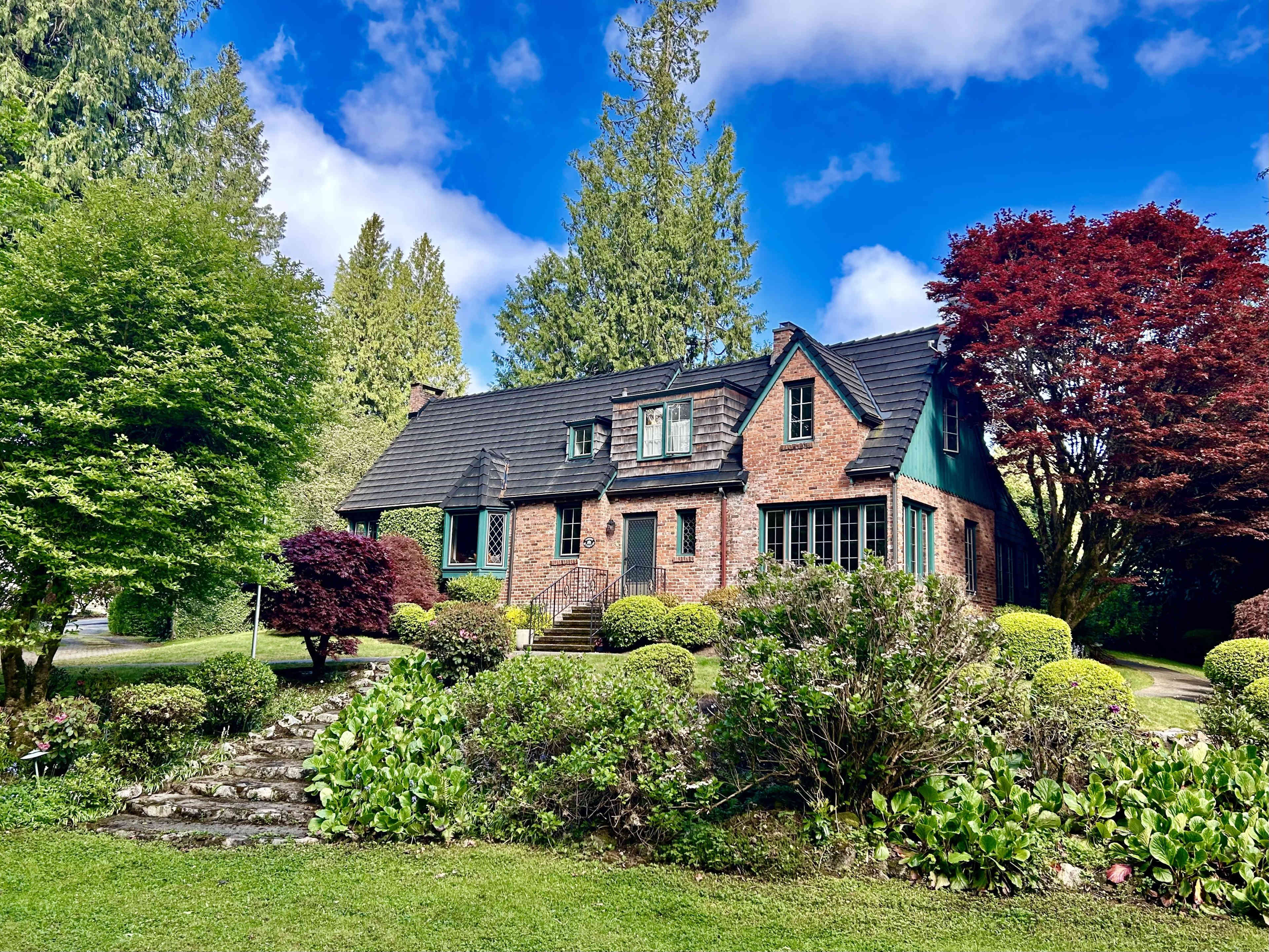 A brick house with a steep roof is surrounded by neatly trimmed bushes and trees under a blue sky with scattered clouds.