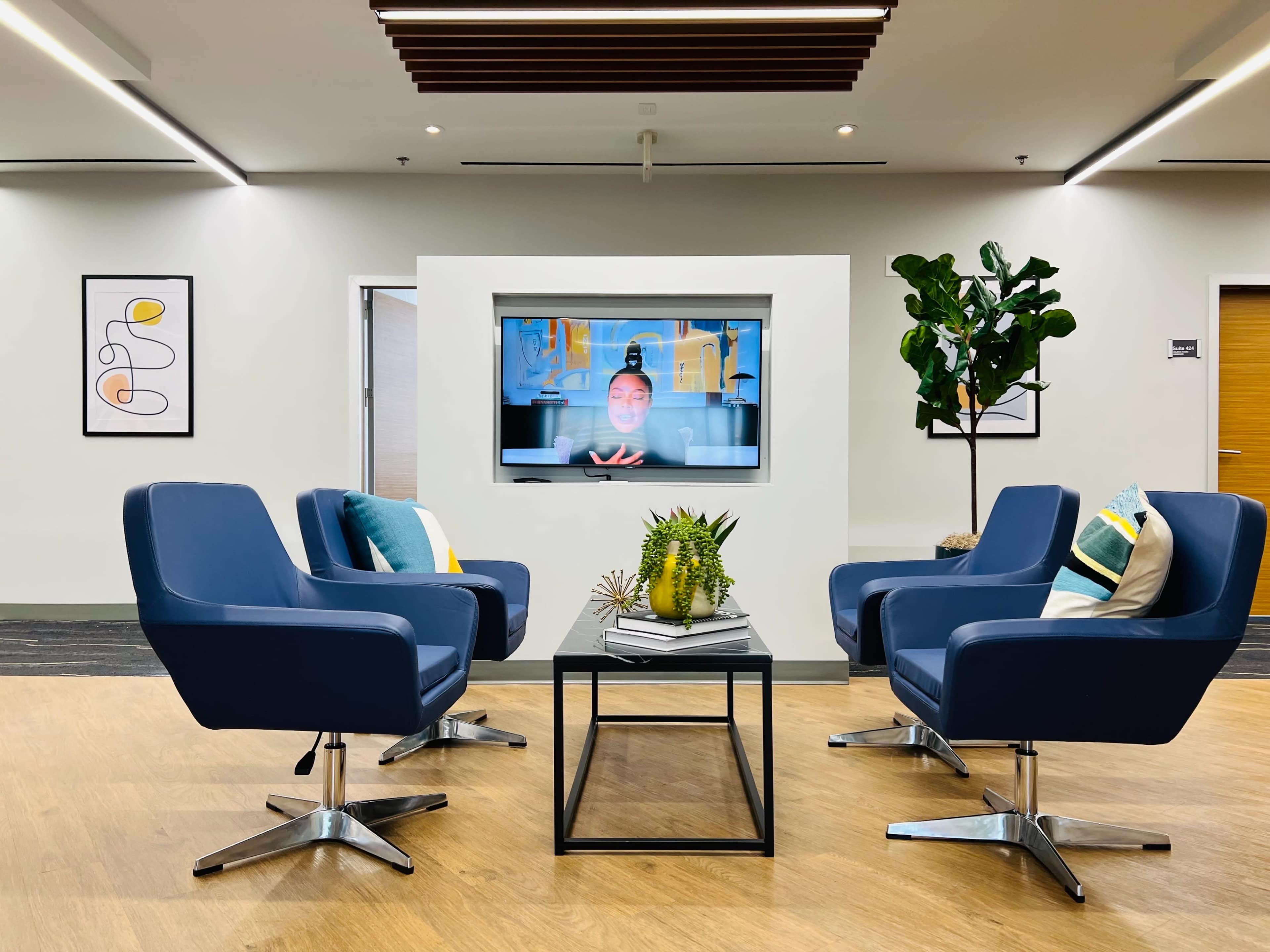 A modern lounge area features four blue chairs arranged around a glass coffee table, with a large television displaying a person, and potted plants in the background.
