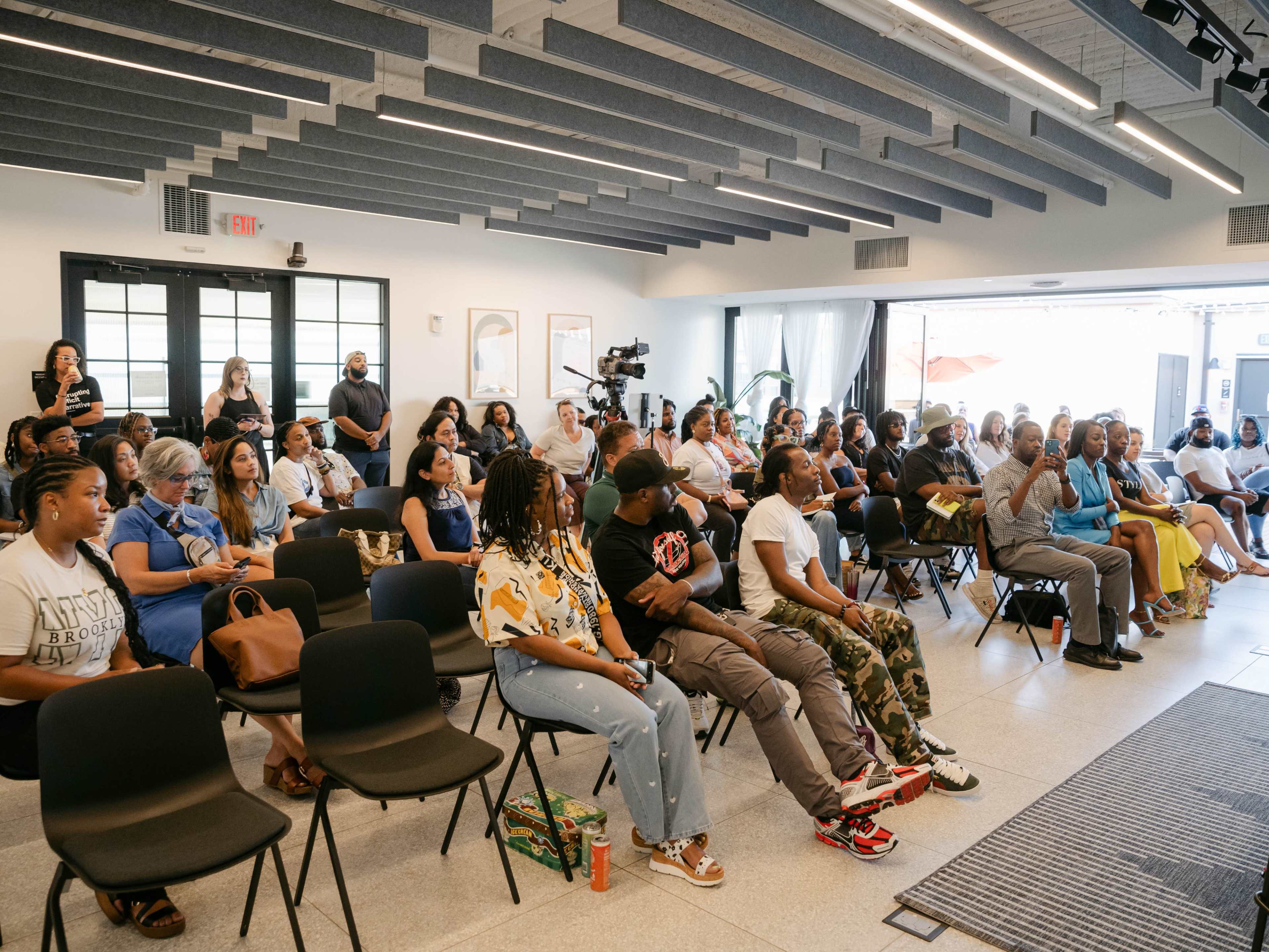 A diverse group of people sits in chairs facing a speaker at a well-lit indoor event.