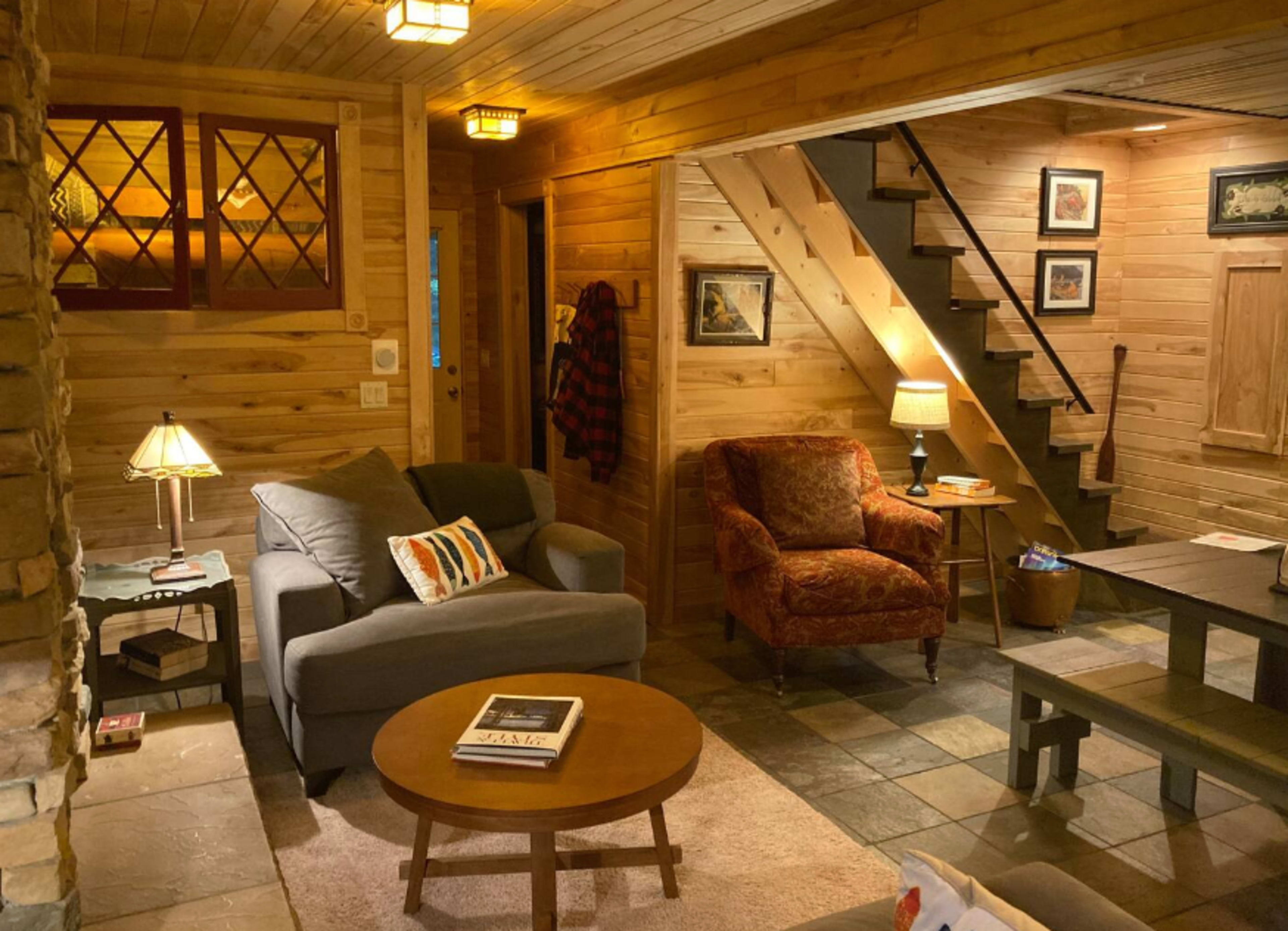 The interior of a wooden-paneled living room features a gray sofa, an ornate armchair, a round coffee table, and a staircase leading to the upper floor.