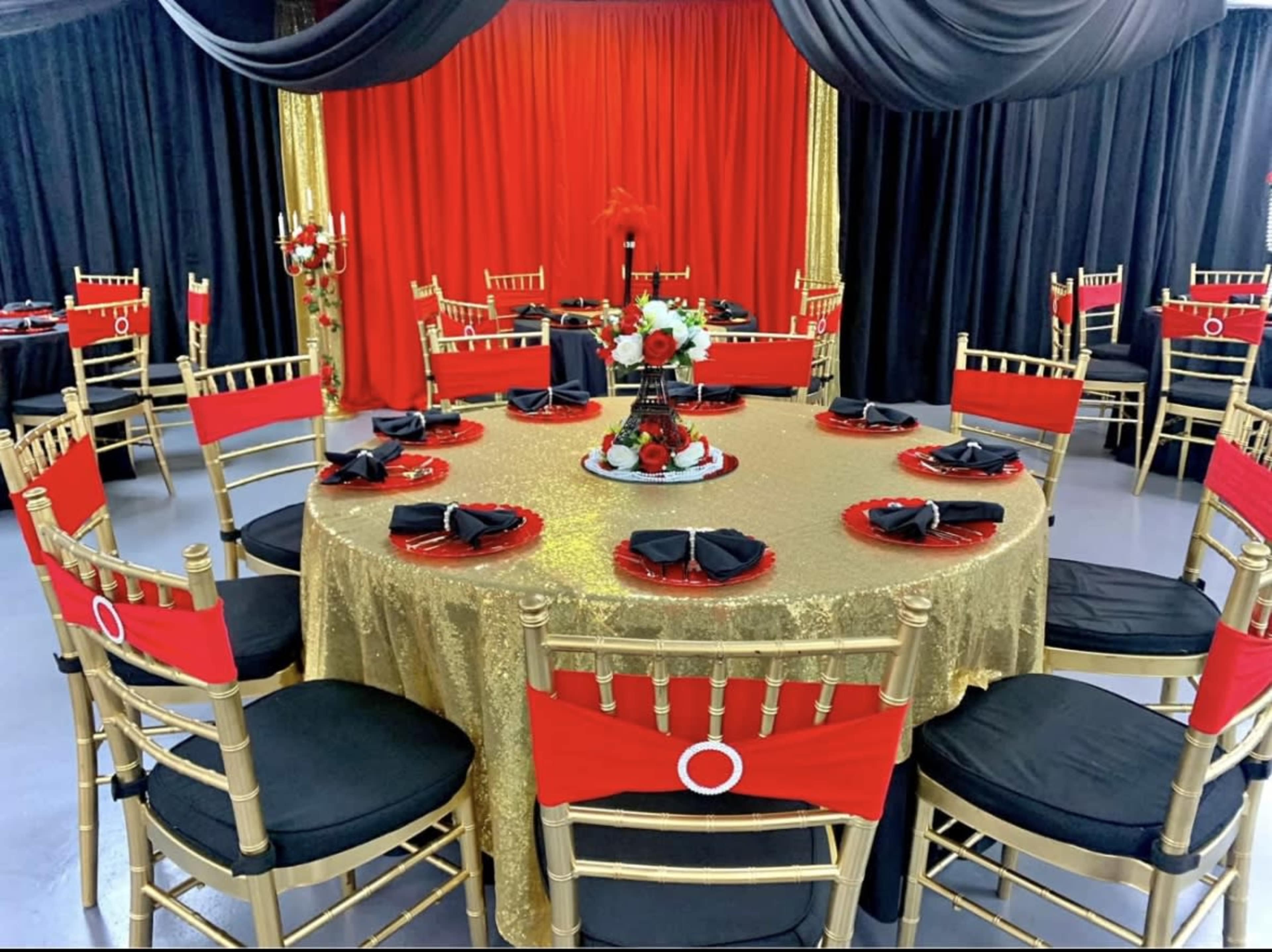 The image shows a banquet table set for a formal event, featuring a golden tablecloth with red and black decorations, gold chairs with red seat covers, and centerpieces adorned with flowers.
