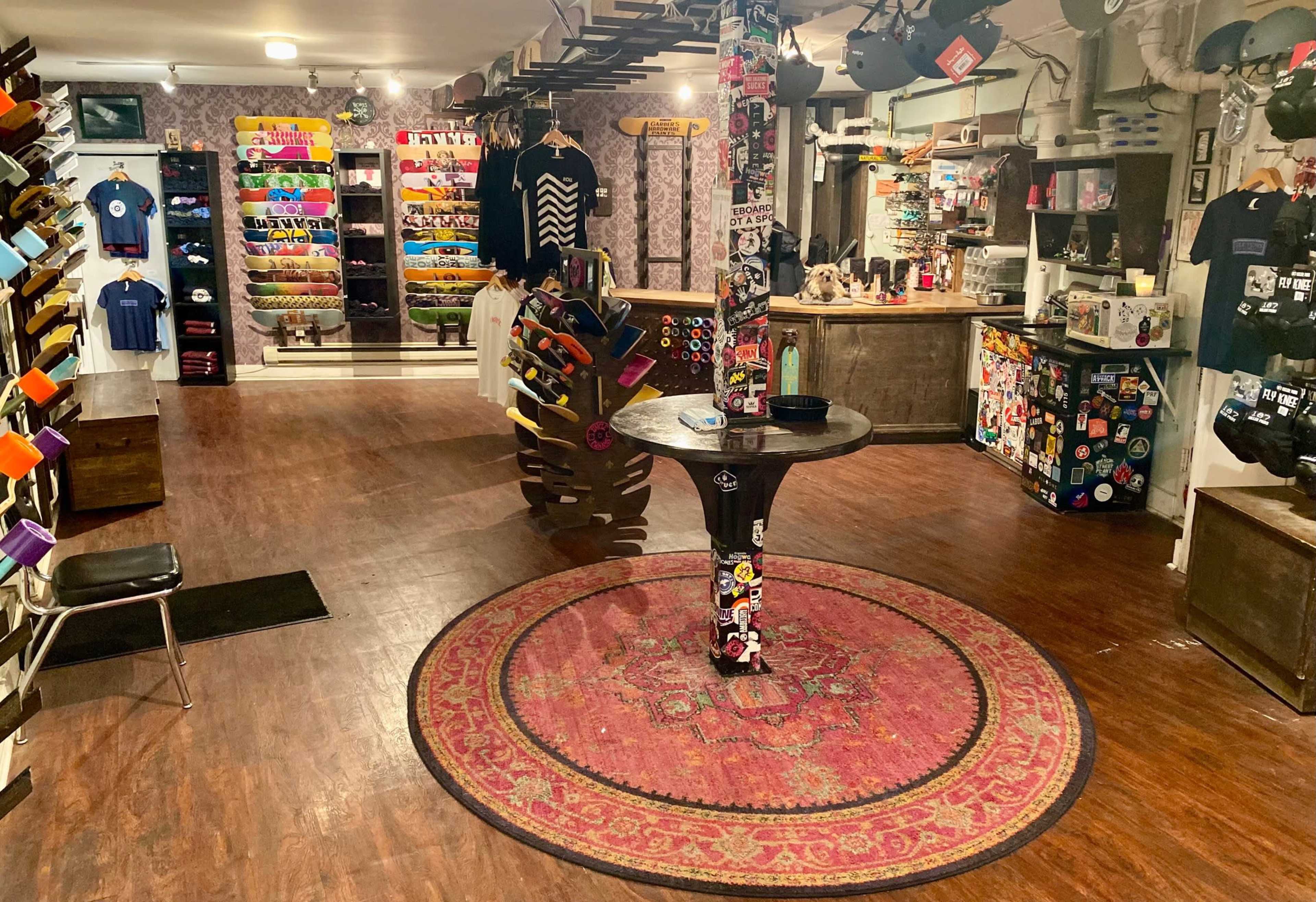 An indoor shop with a wooden floor, featuring a circular rug in the center surrounded by shelves displaying various skateboards, clothing, and accessories.