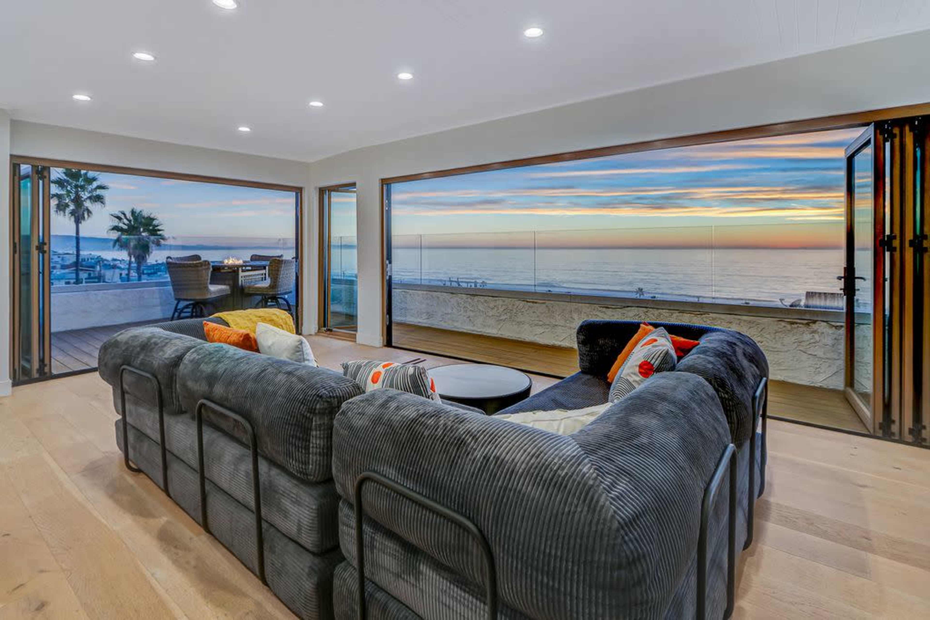 The image shows a modern living room with large windows offering a view of the ocean and a sunset, featuring a cozy sectional sofa and a circular coffee table.