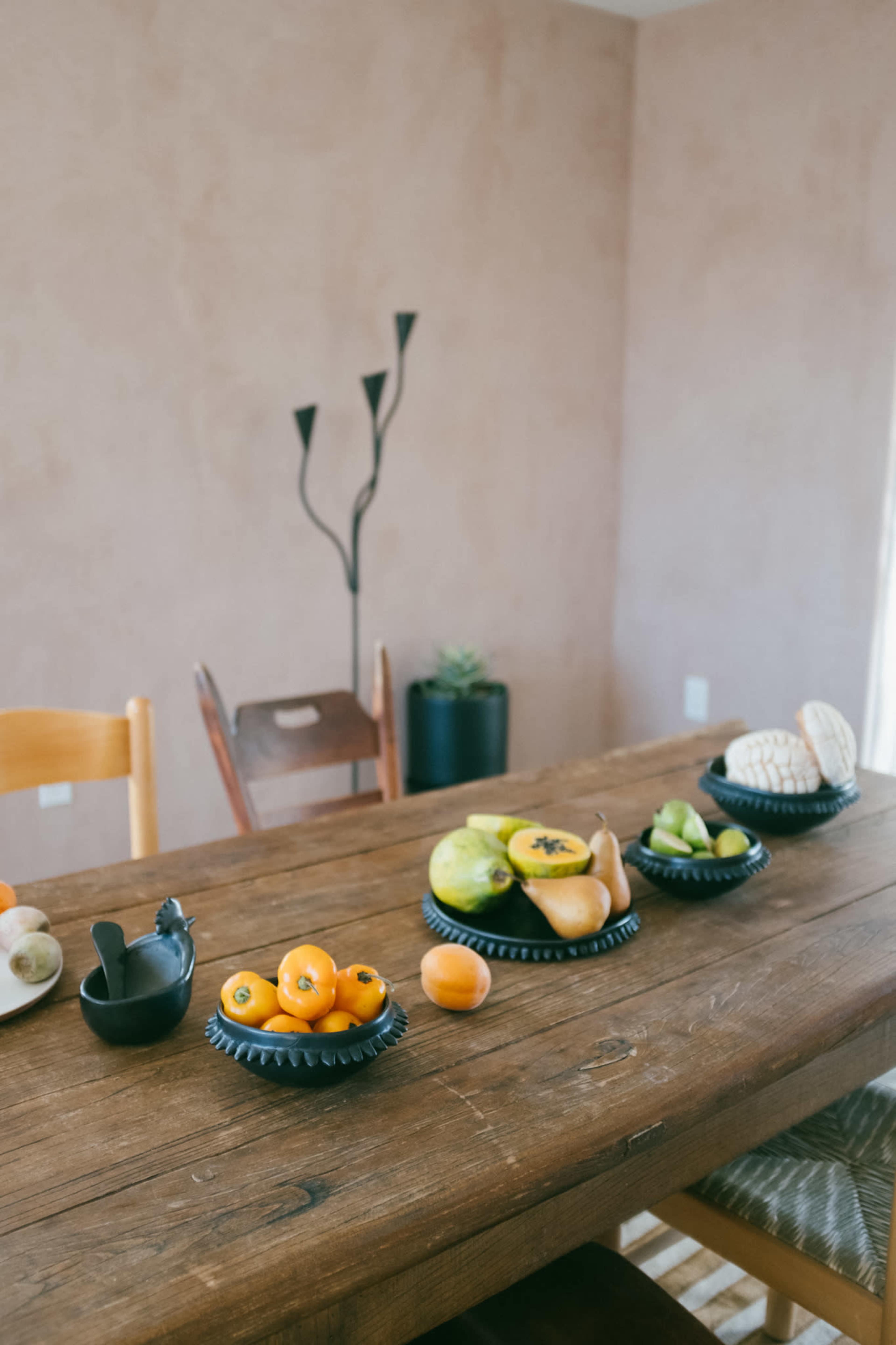 A wooden table is set with several bowls of assorted fruits and a decorative candle holder in a minimalistic room.