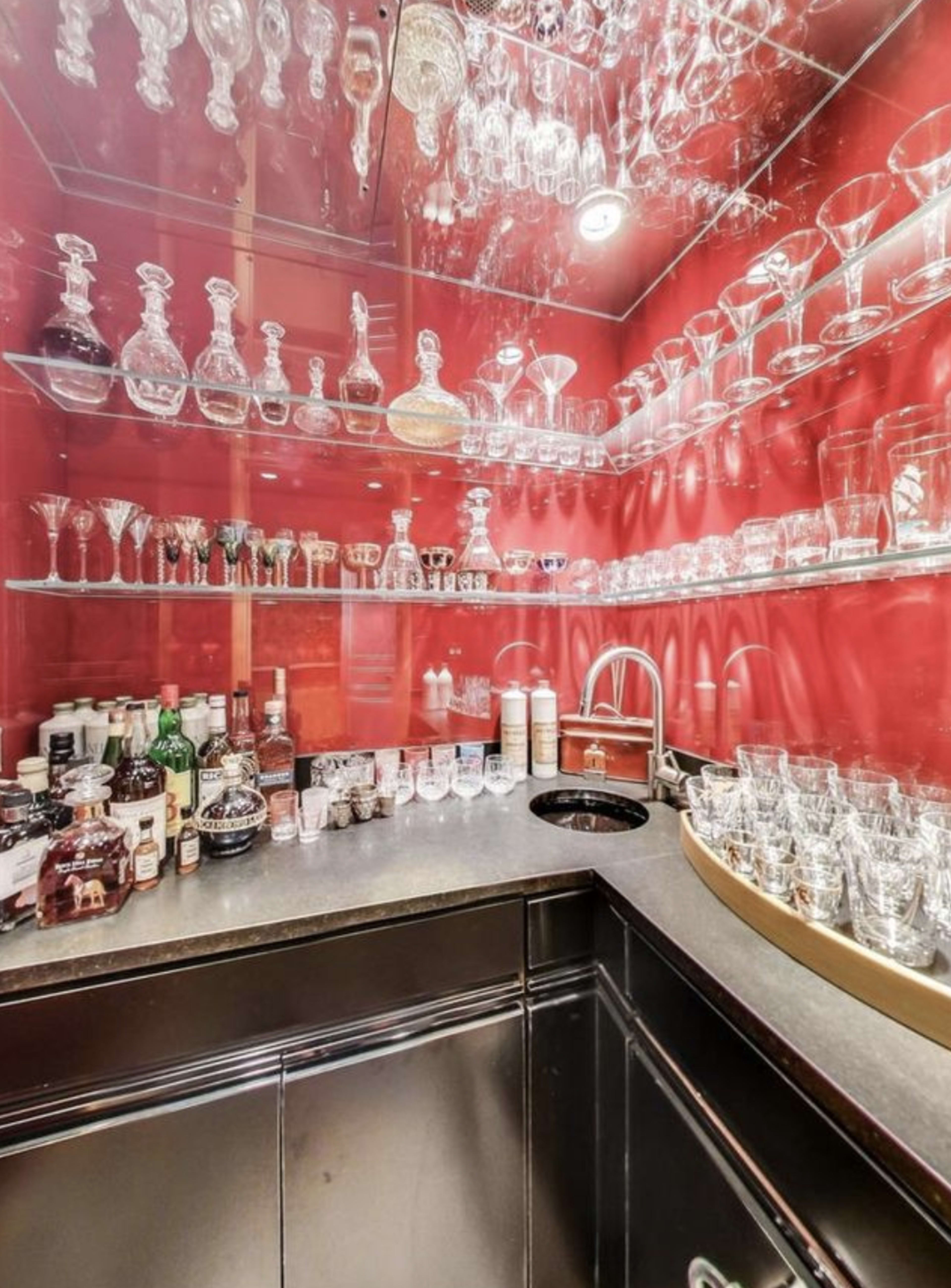 A well-organized home bar features a variety of glassware and liquor bottles displayed on shelves against a red backdrop.
