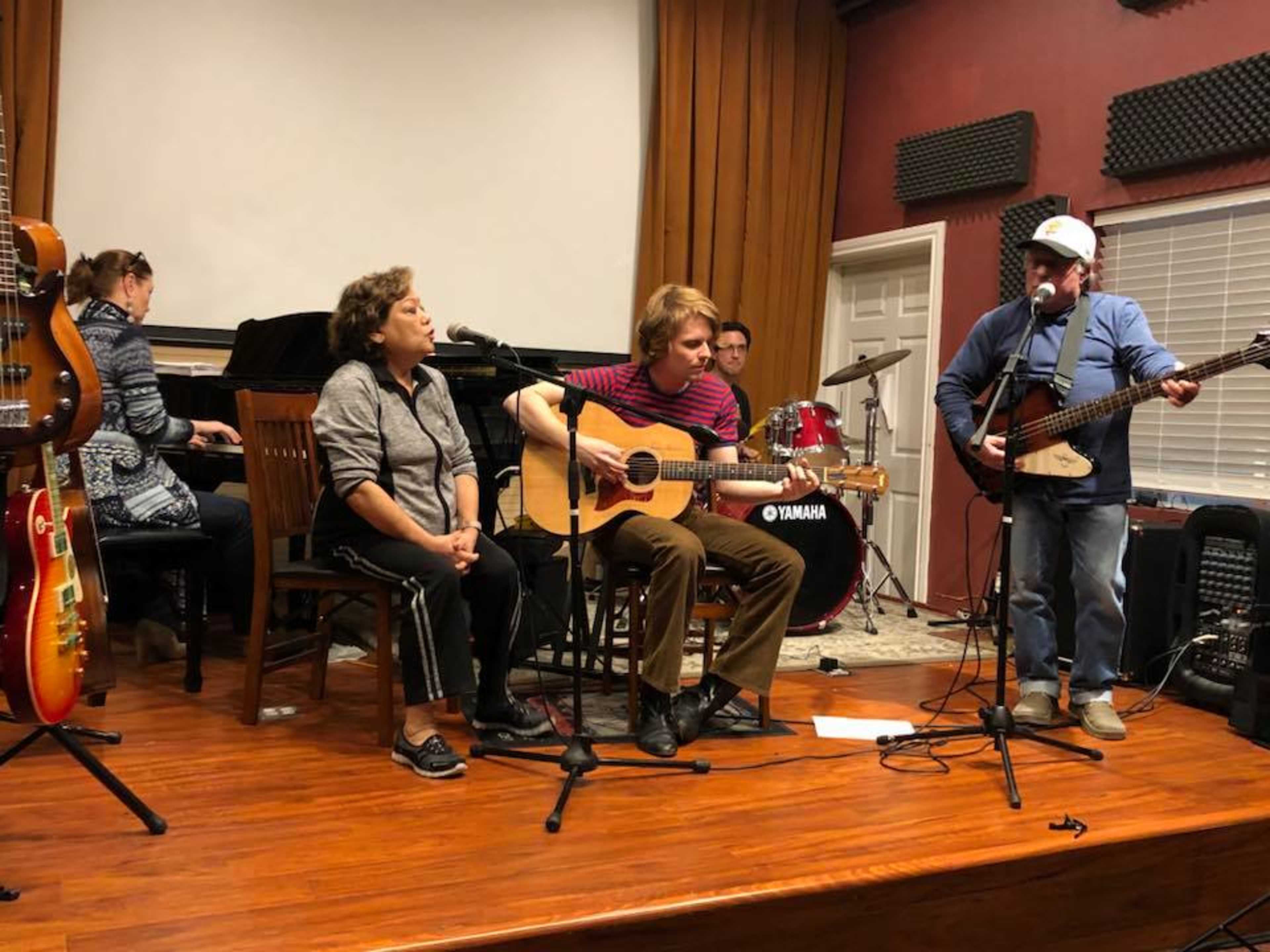 A group of musicians is performing a live session, featuring a singer with an acoustic guitar, a woman seated beside him, and two other musicians playing a guitar and drums in the background.