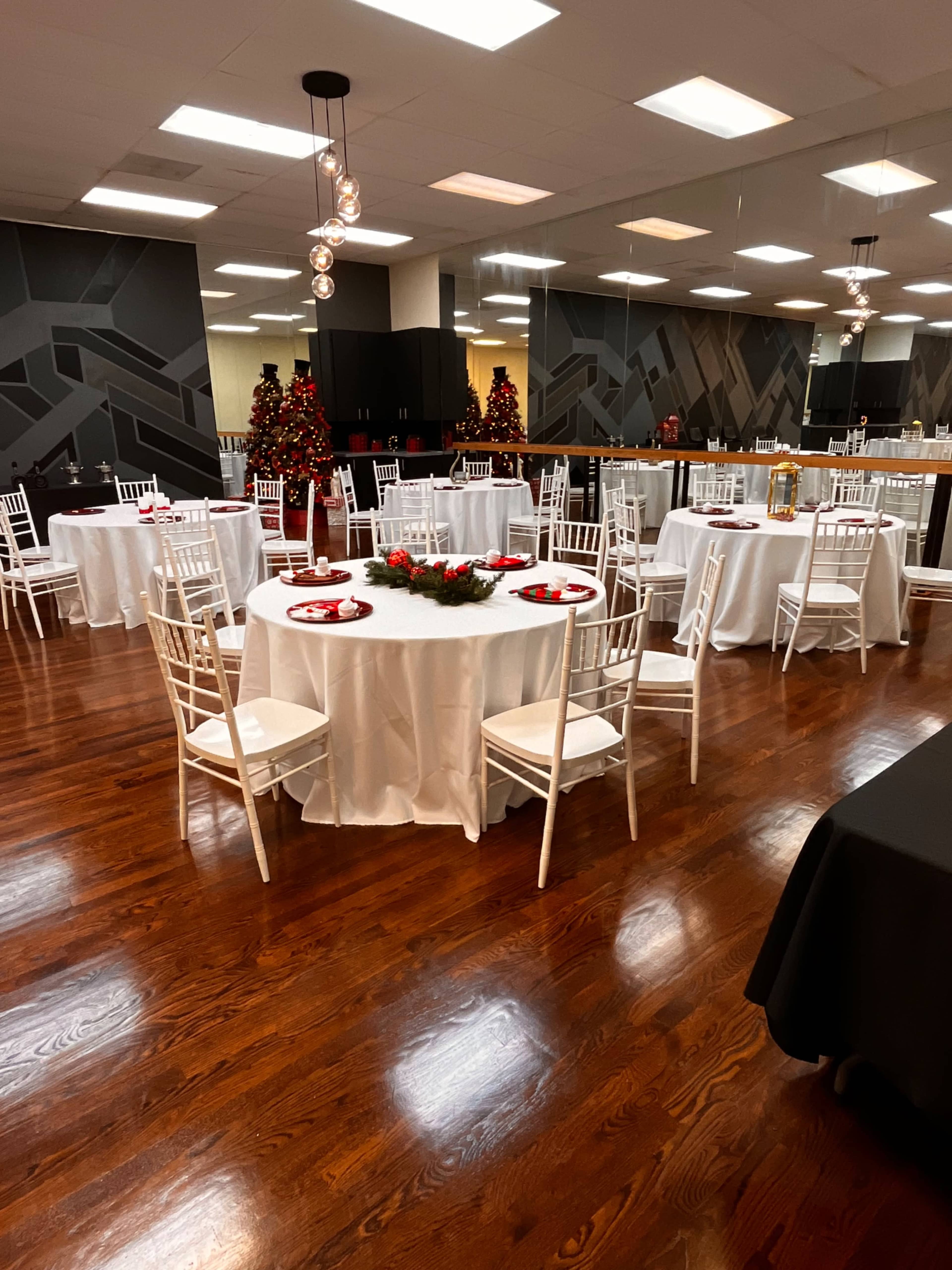 The image shows an event space arranged with several round tables covered in white linens, decorated for a festive occasion with red accents and Christmas trees in the background.