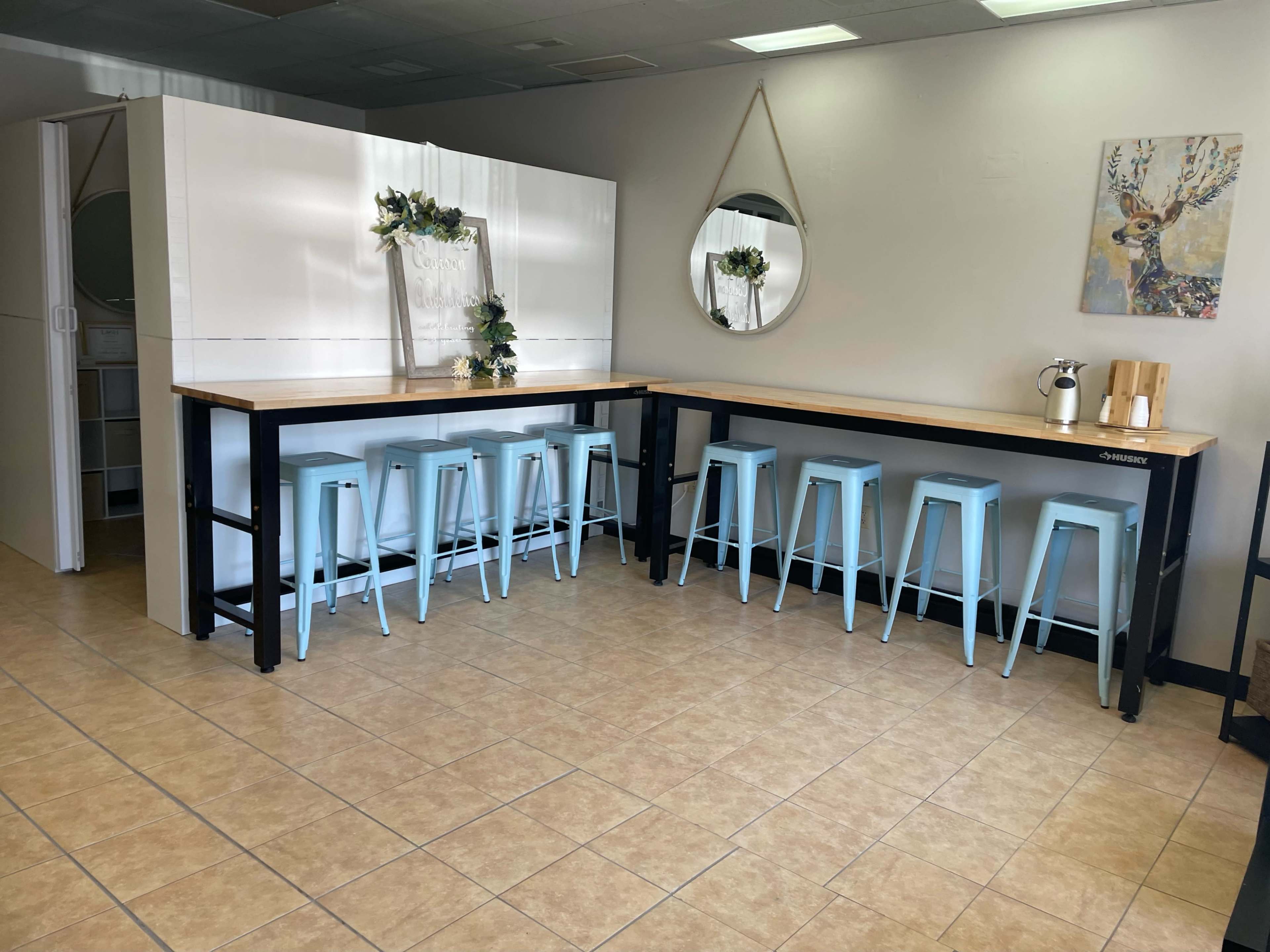 The image shows a bright, minimalistic interior featuring two long tables with light blue stools and a round mirror on the wall.