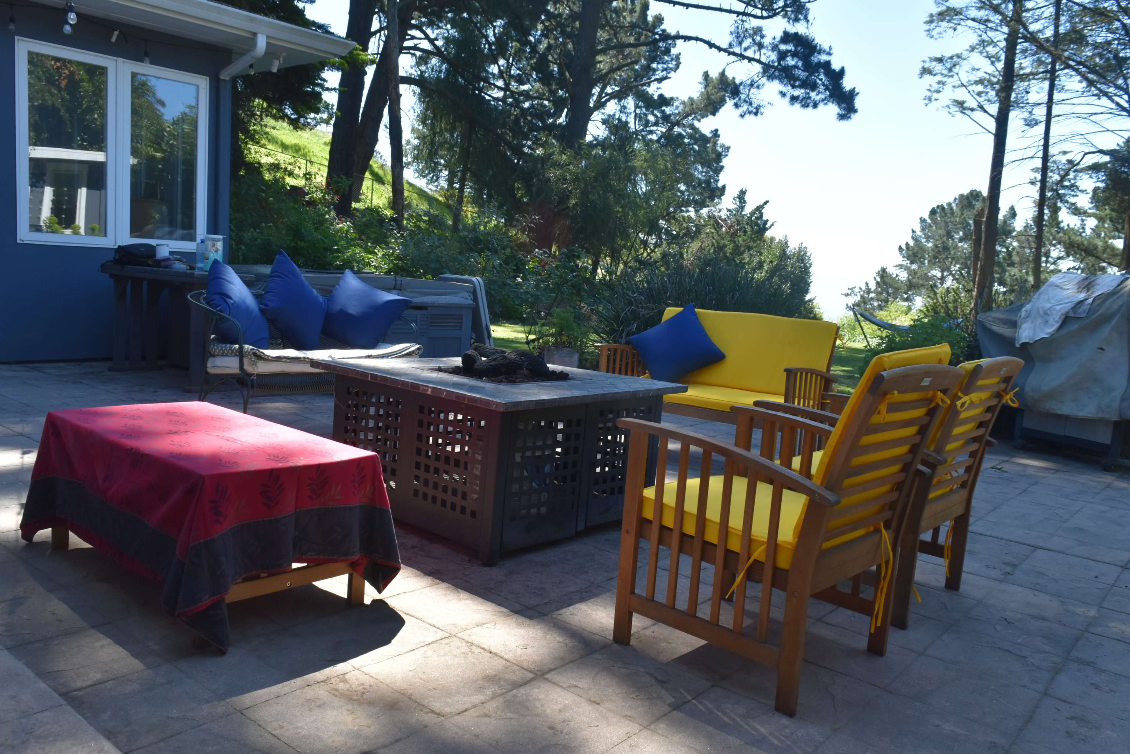 The scene features an outdoor patio with wooden seating, a low table, and scattered cushions, surrounded by trees and greenery.