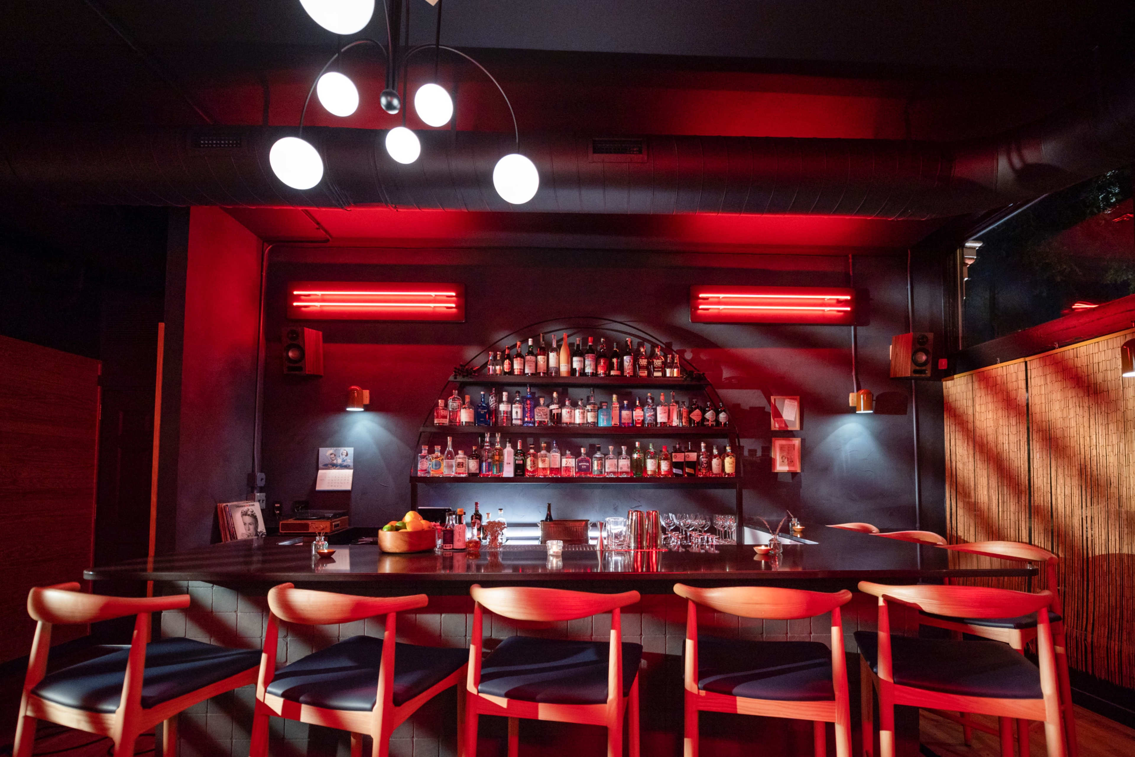 A modern bar scene features a long wooden counter with empty chairs, illuminated by red lights, showcasing a wall of various bottles in the background.