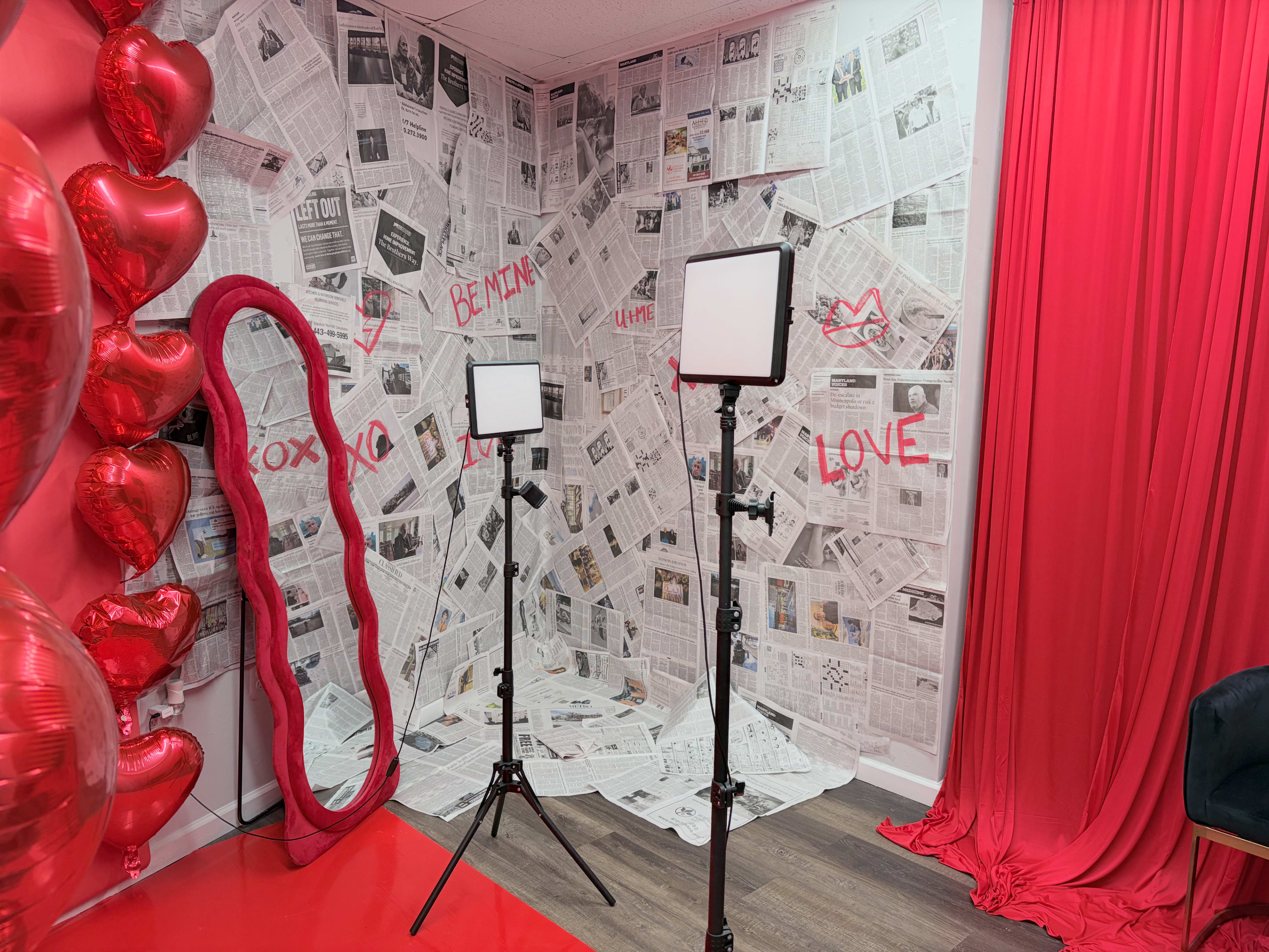 The image shows a photo booth setup with a wall covered in newspaper clippings, two light stands, red fabric draping, and heart-shaped balloons.