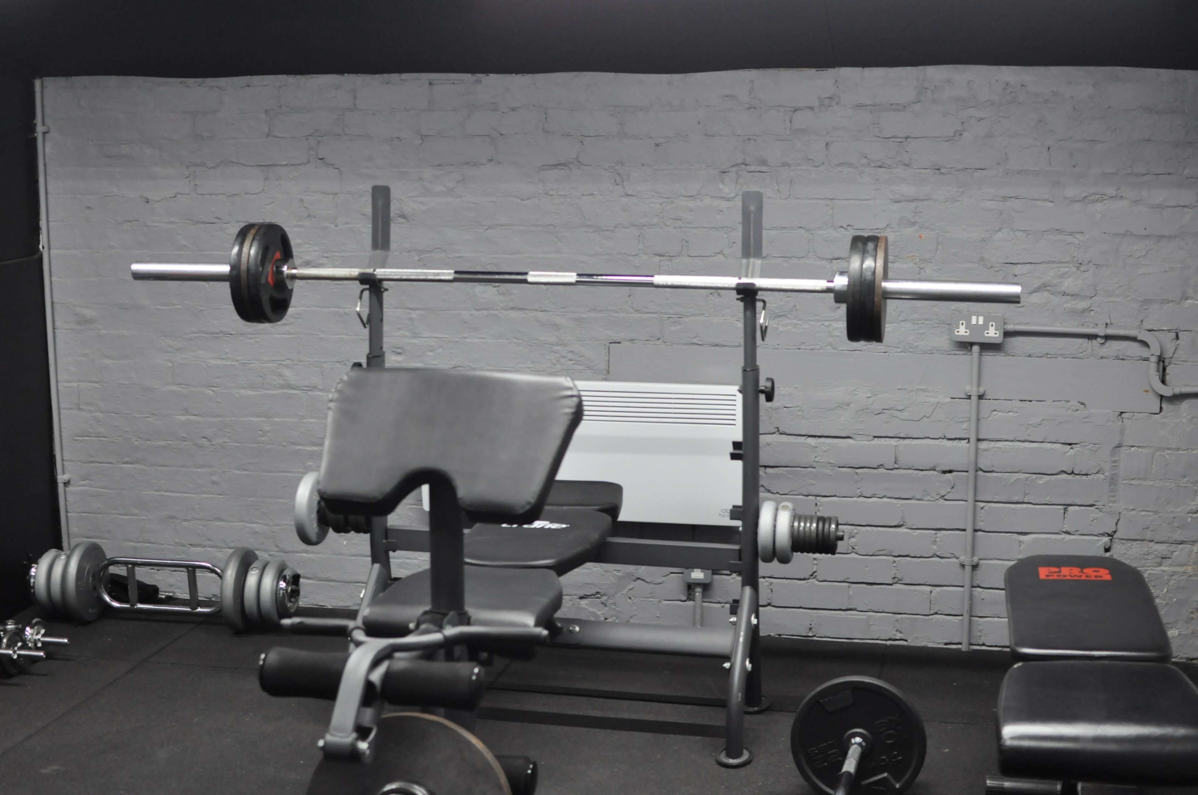 The image shows a weight bench with a barbell loaded with weight plates and an exercise area equipped with various gym equipment.