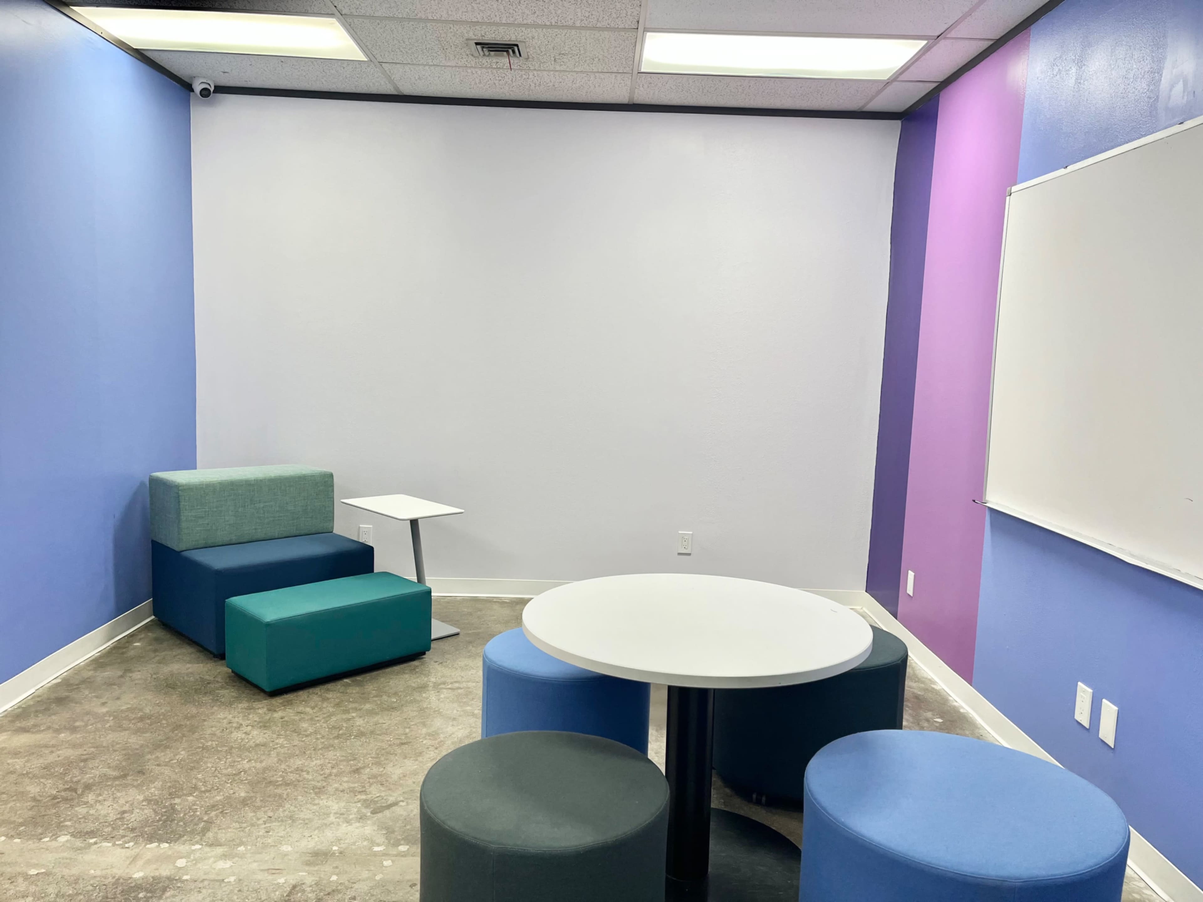 The image shows a small, minimalistic room with colorful walls and furniture, featuring a whiteboard, a round table, and several seating components.