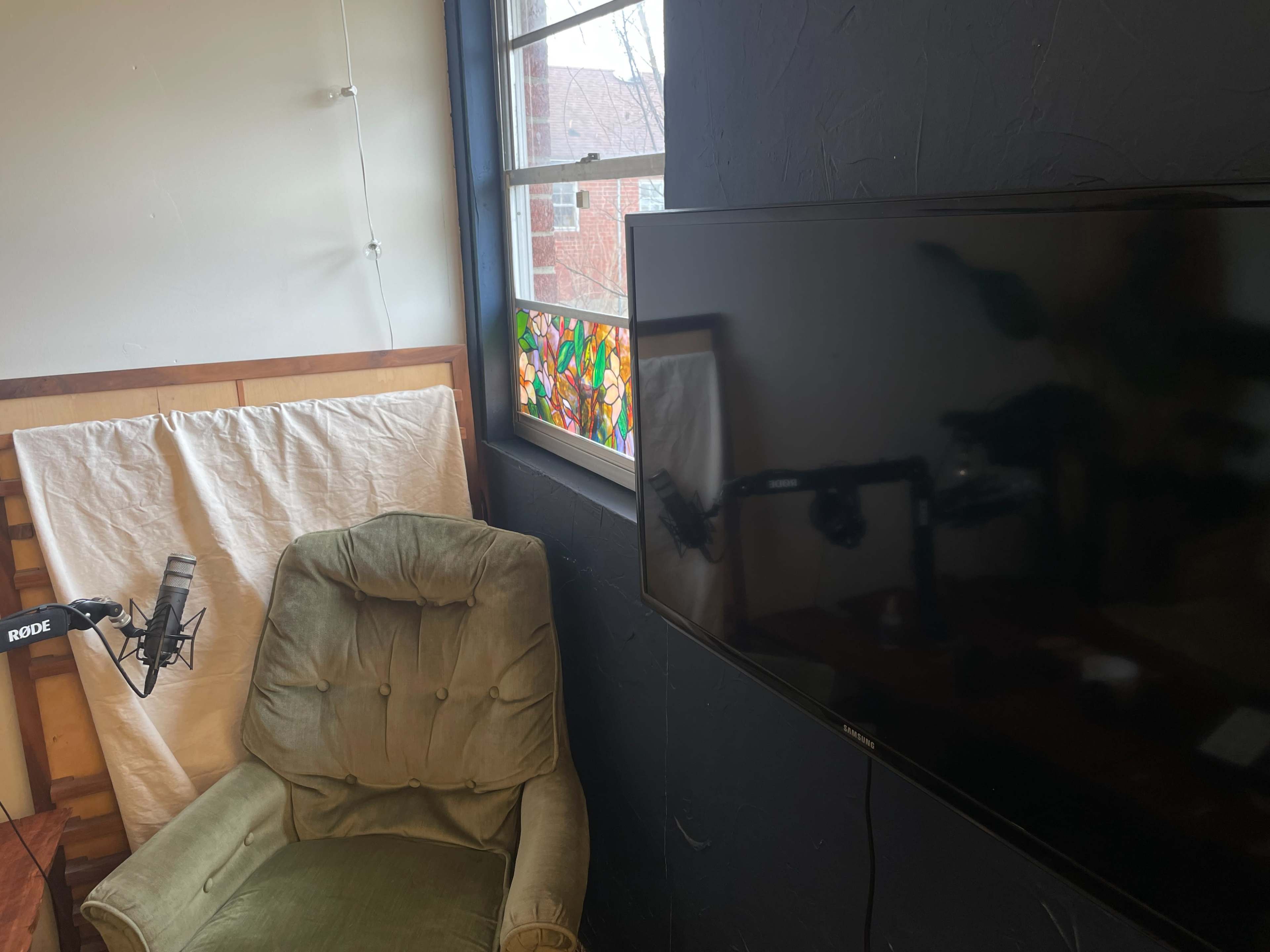 The image shows a corner of a room featuring a green upholstered armchair, a microphone on a stand, and a television next to a colorful stained glass window.