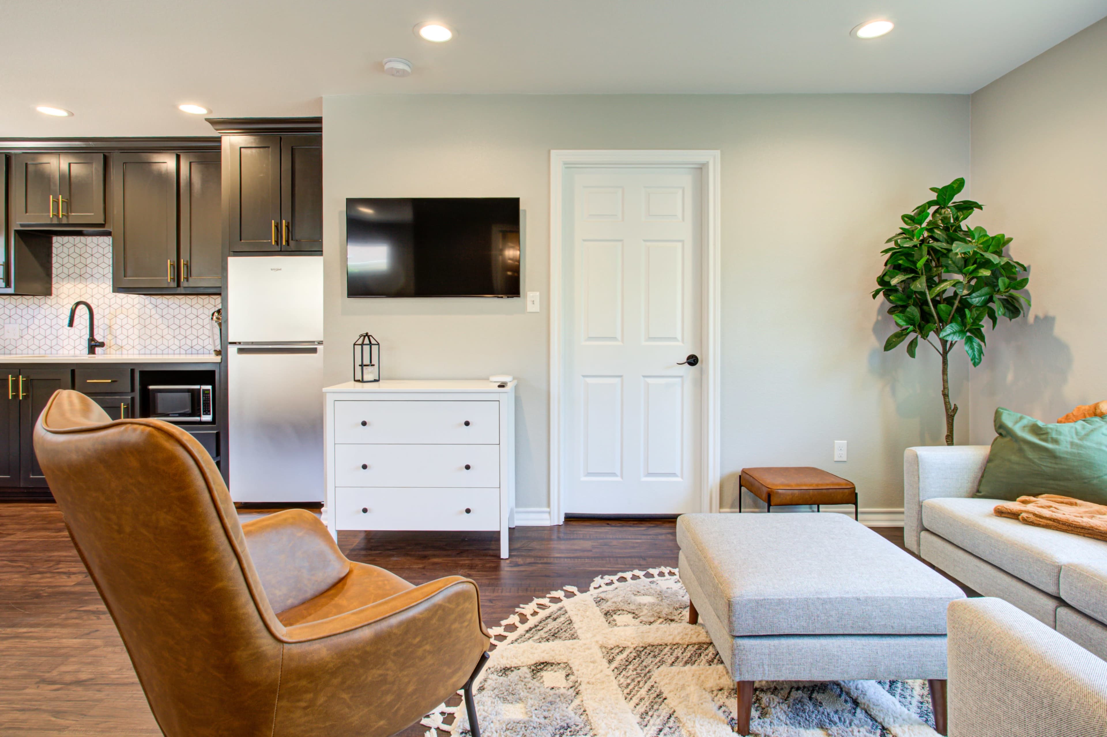 A modern living room with a brown armchair, a light gray sofa, a white dresser, and a plant, all set against a backdrop of dark kitchen cabinetry and a wall-mounted TV.