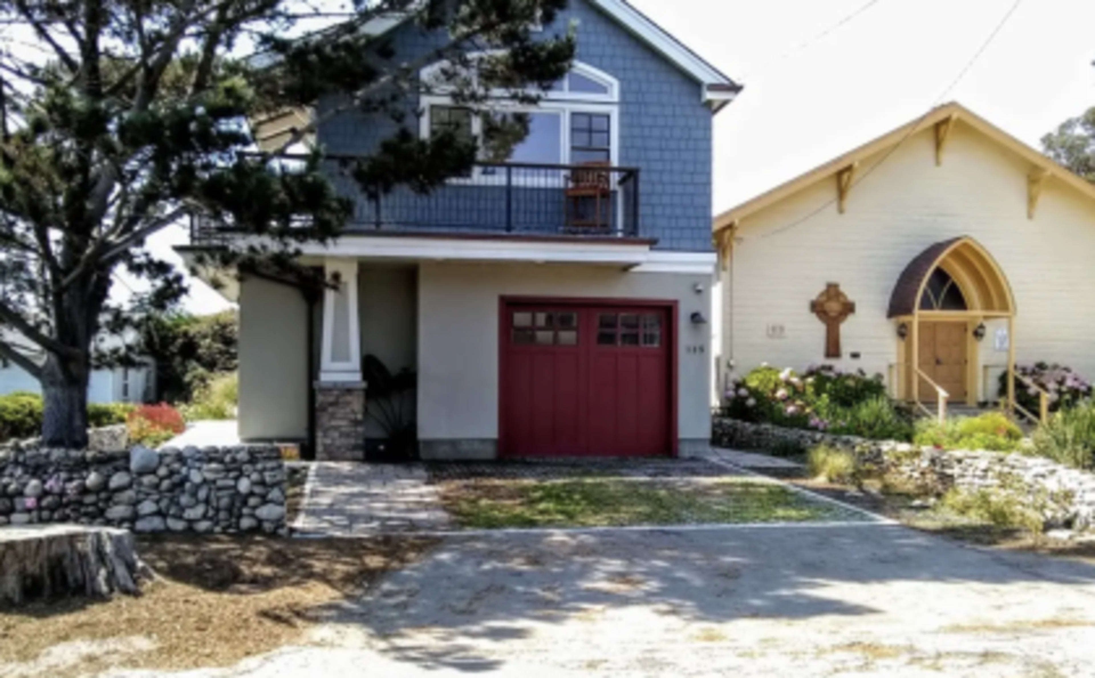 The image shows a residential house with a blue exterior, a red garage door, and an adjacent light-colored building with a church-like architecture.