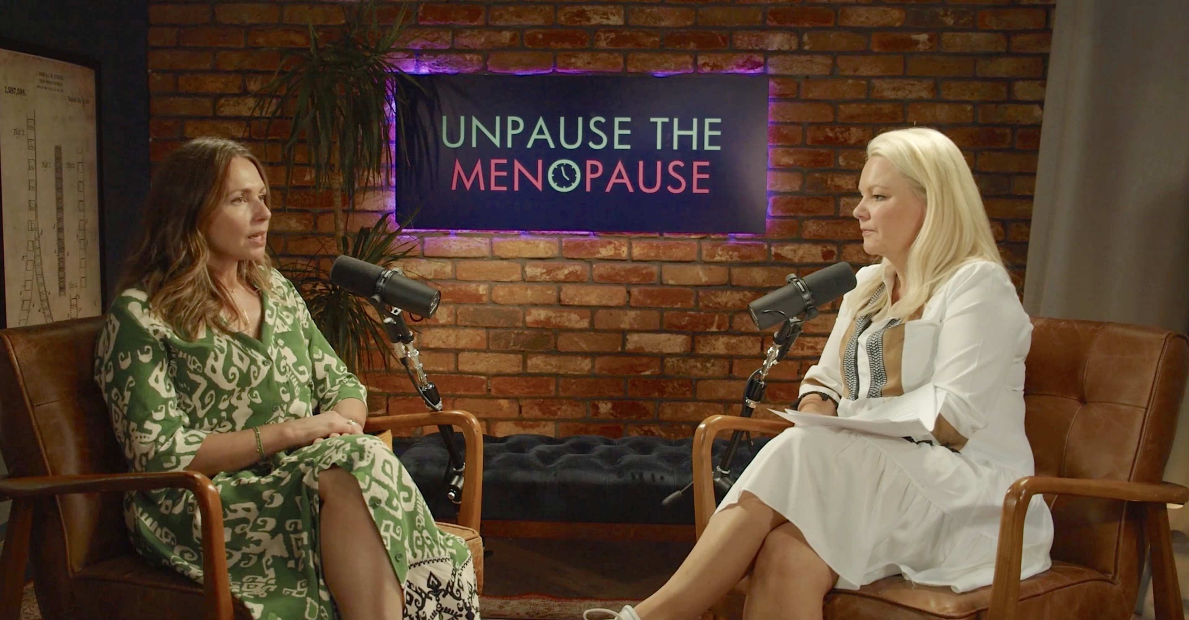 Two women sit in modern chairs in front of a brick wall with a neon sign that reads "UNPAUSE THE MENOPAUSE," each positioned with a microphone in front of them.
