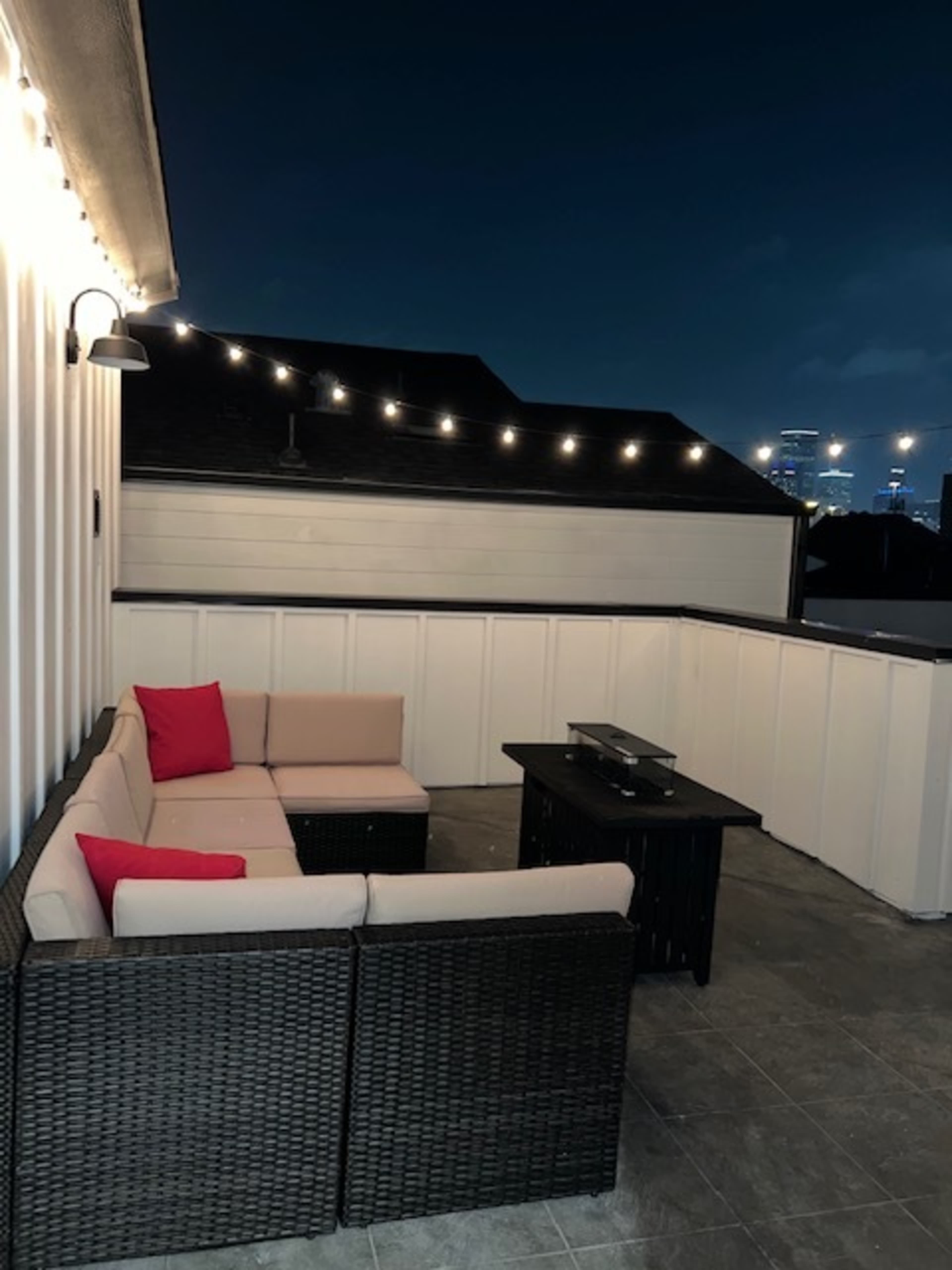 A rooftop seating area features a sectional couch with red accent pillows and a small coffee table, illuminated by string lights against a night sky.