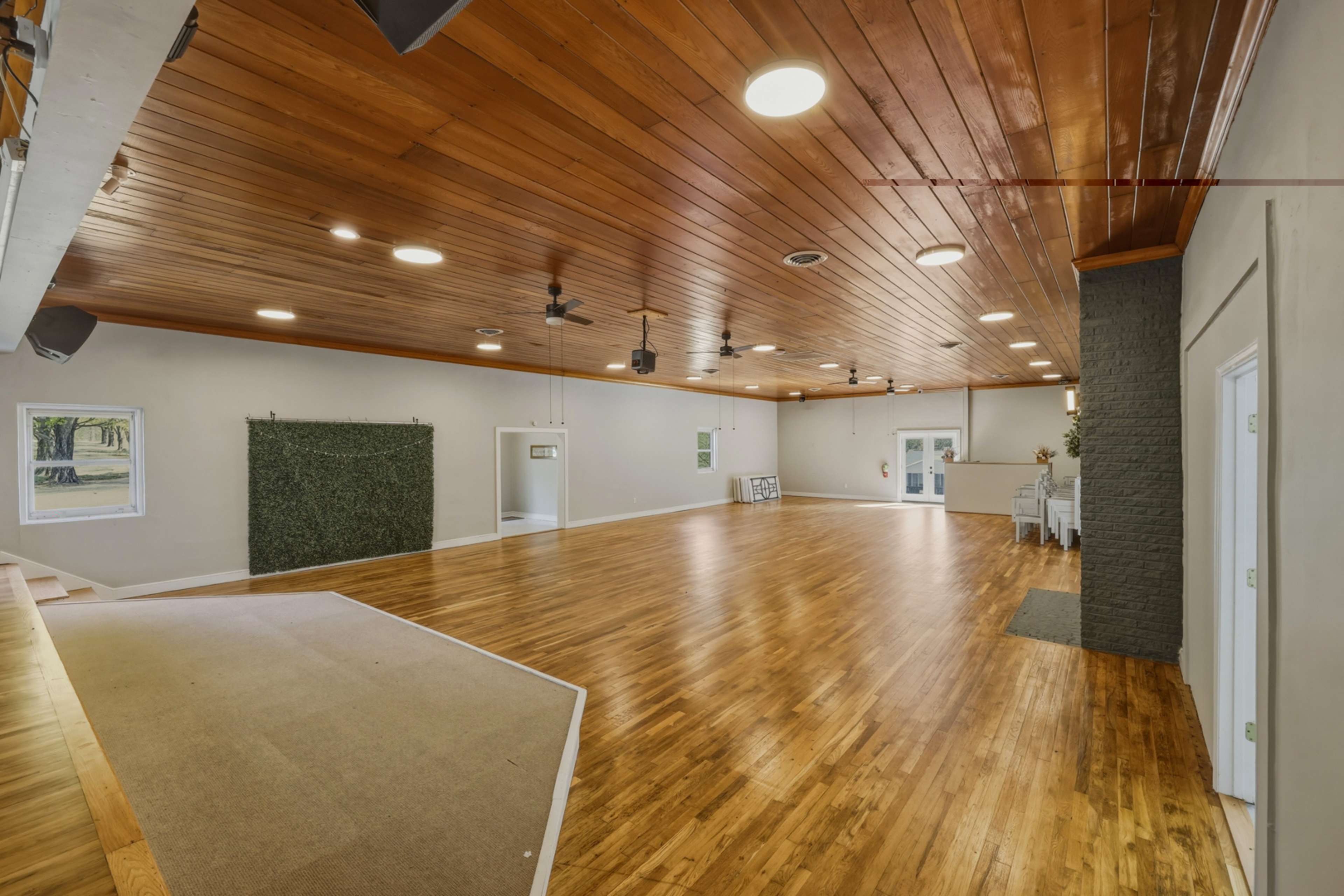 The image shows a spacious, empty indoor venue with wooden flooring, a wooden ceiling, and a section featuring a green wall.