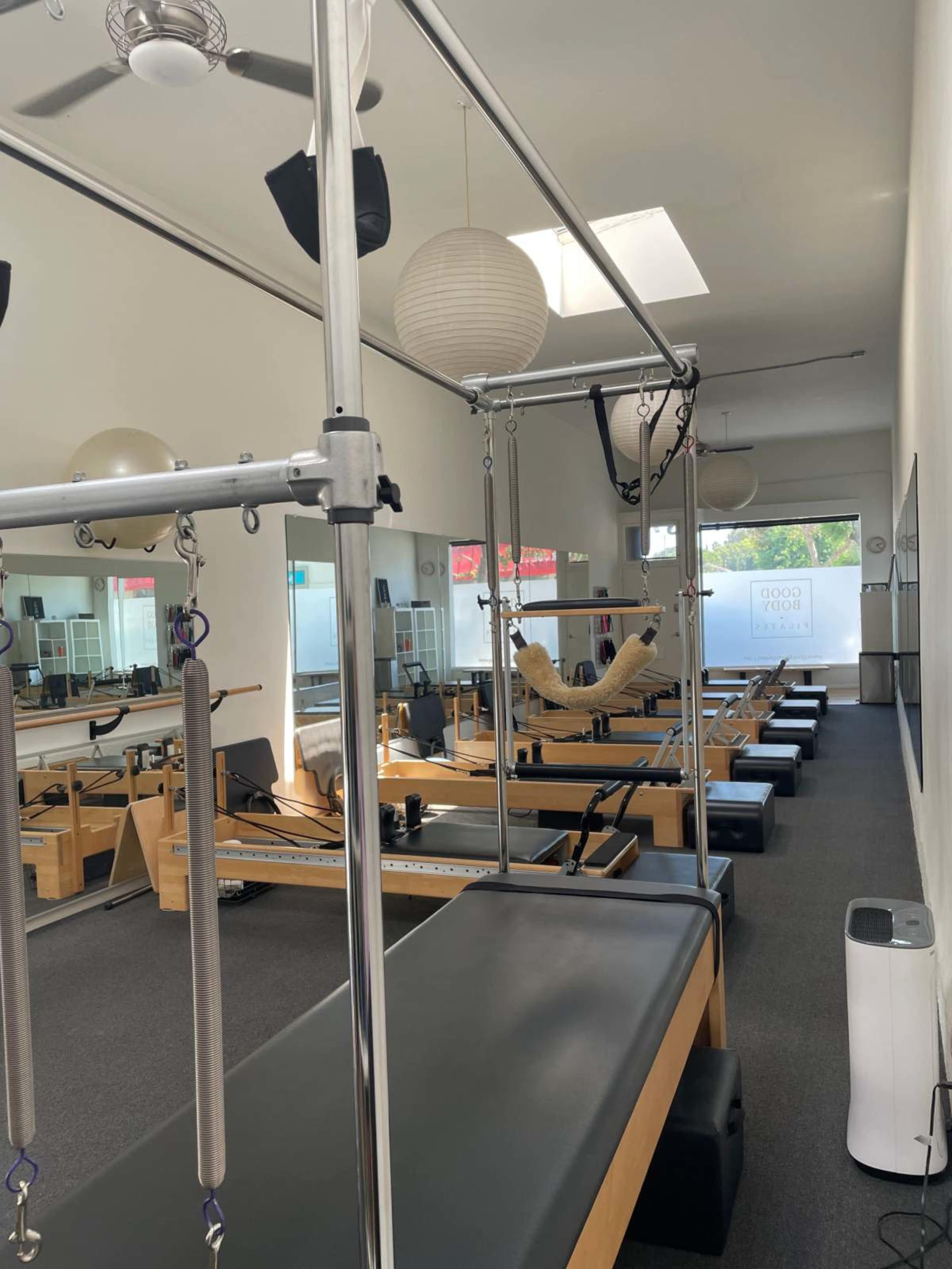 The image shows a Pilates studio with reformer machines arranged in rows beneath large mirrors and bright lighting.