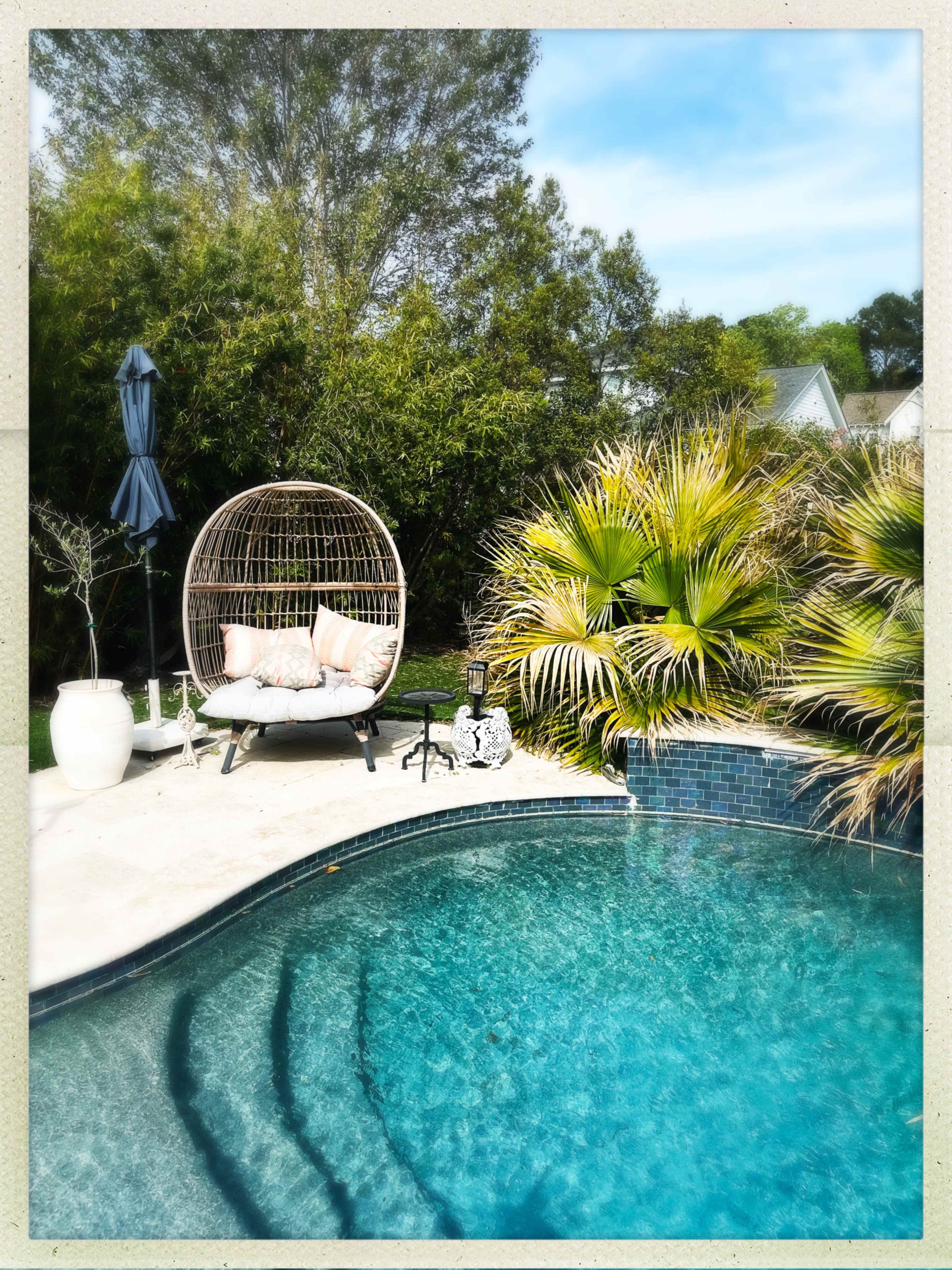 A cozy round chair sits beside a clear swimming pool, surrounded by lush greenery and landscaping.