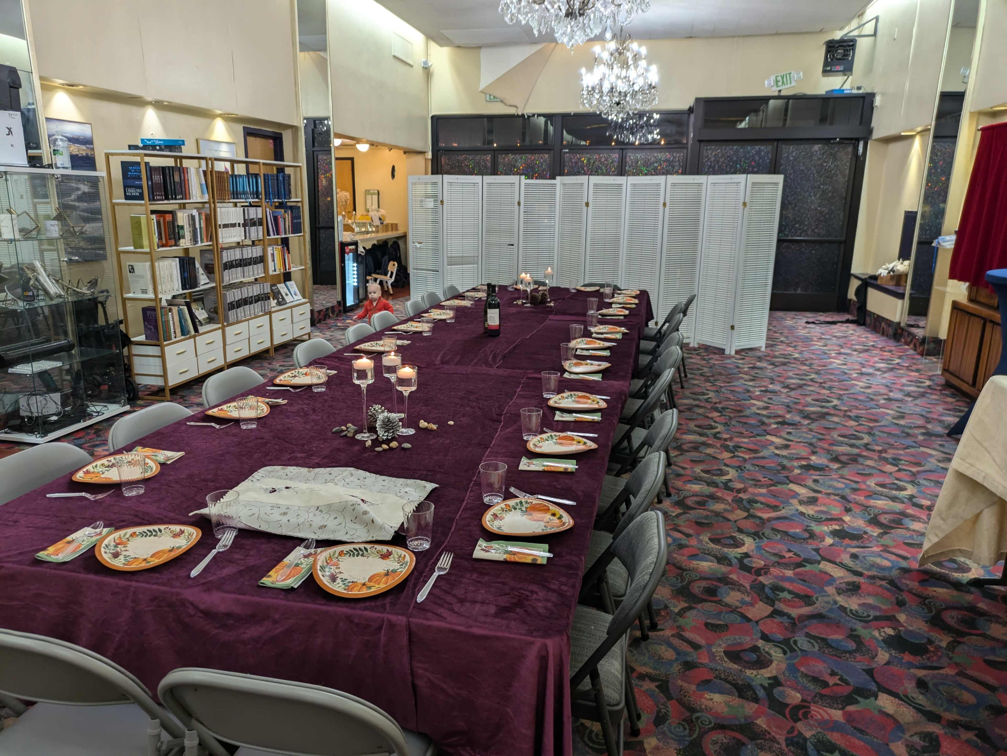 A long dining table is set with plates and glassware, surrounded by chairs, in a banquet room adorned with patterned carpeting and a chandelier.
