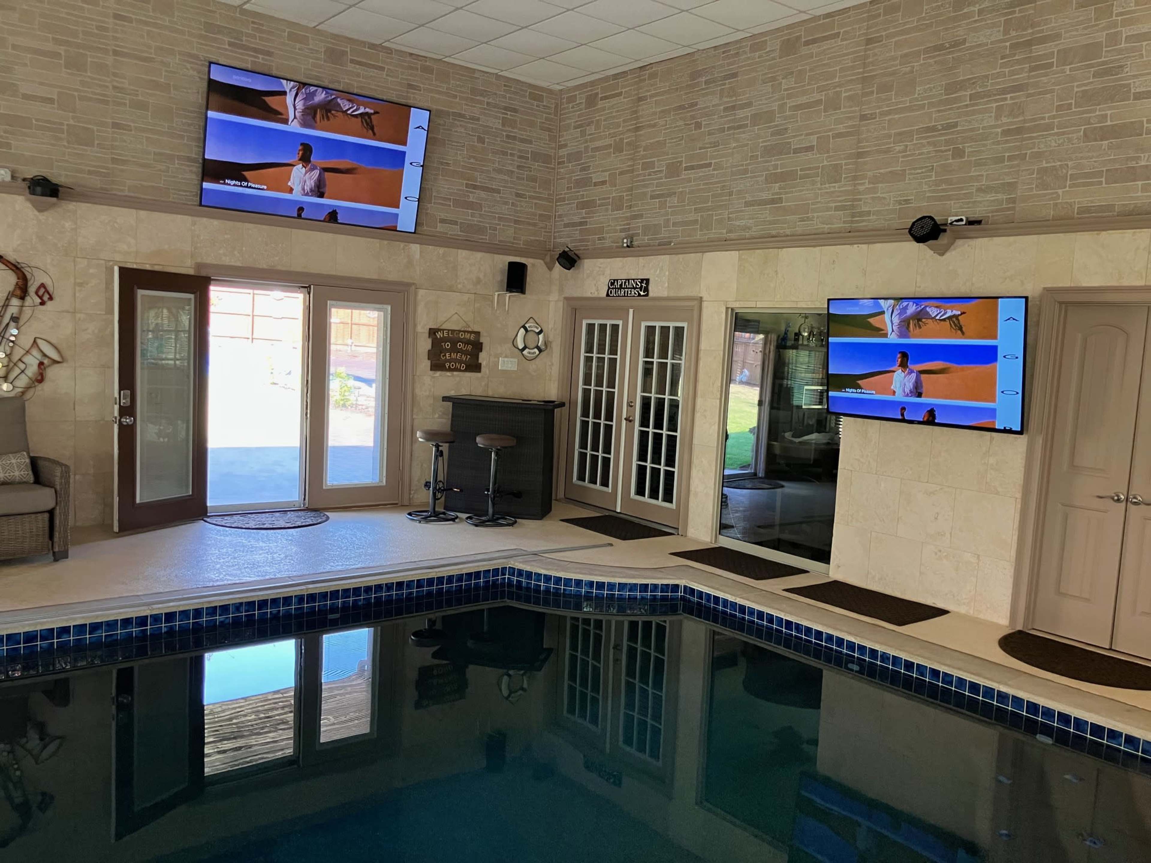 The image shows an indoor pool area with two large screens mounted on the walls, a bar area with stools, and sliding glass doors leading outside.