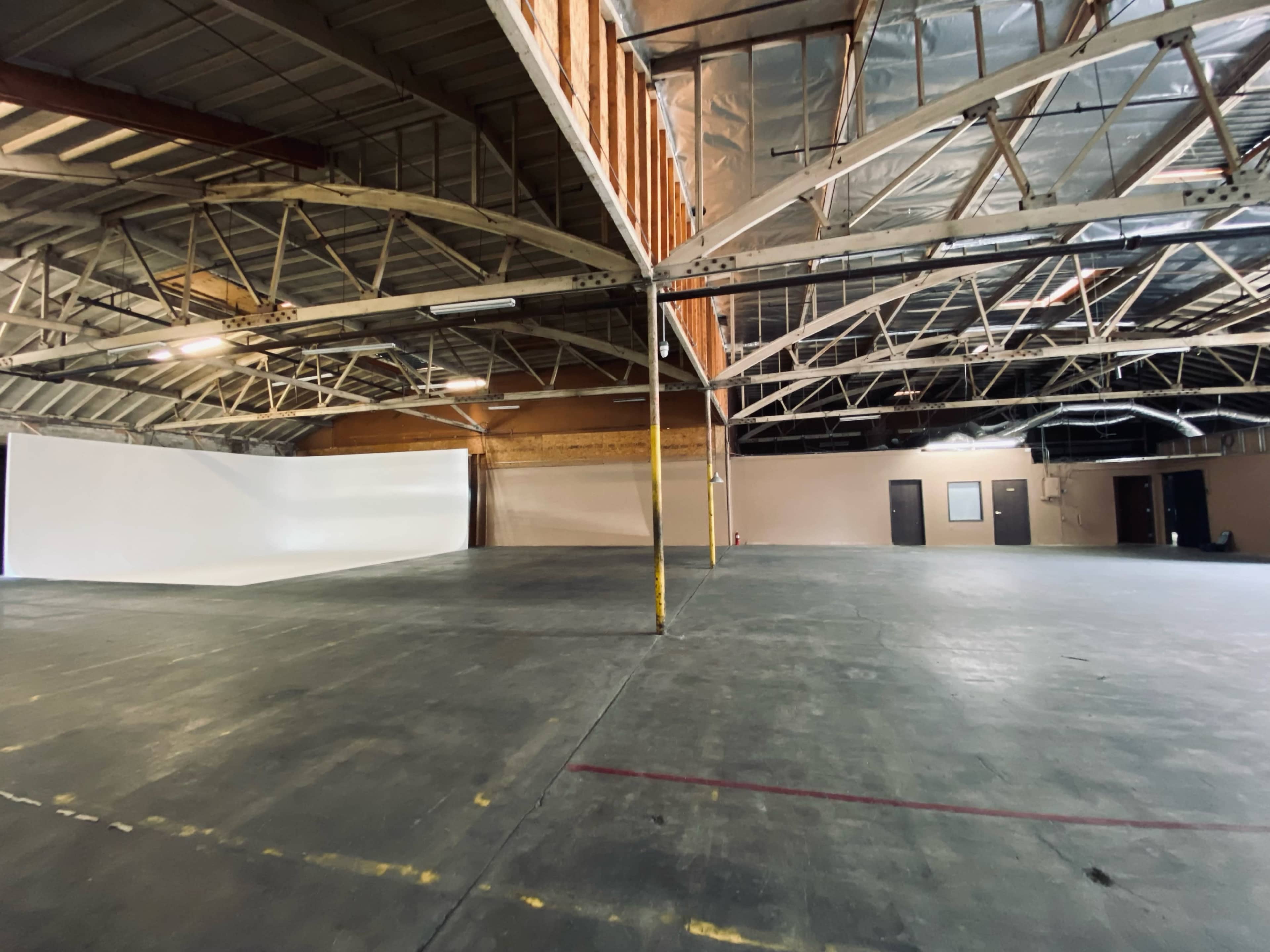 An empty industrial warehouse space with high ceilings and a large, unobstructed area featuring a white backdrop along one wall.
