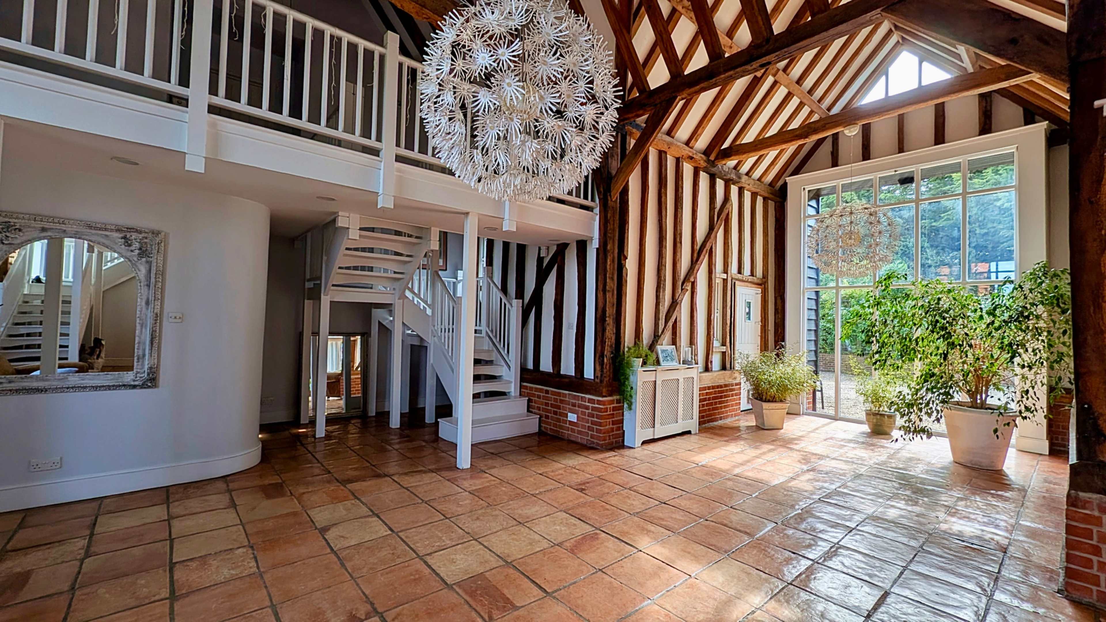 The image shows a spacious, warmly lit interior featuring exposed wooden beams, a spiral staircase, and large windows with potted plants on the floor.